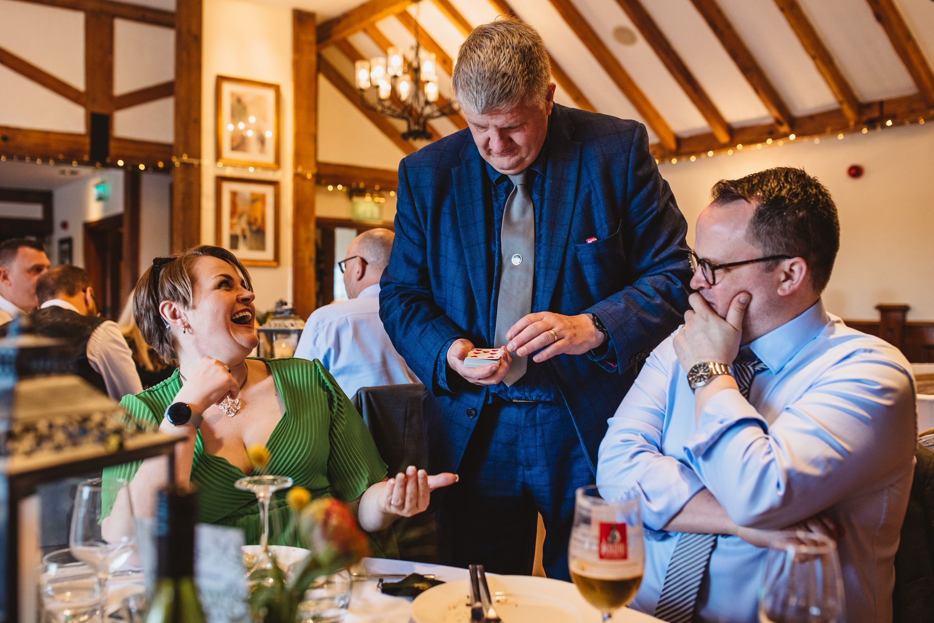 A magician performing a trick at a wedding reception. The guests are amazed and laughing. The scene is indoors, at a table.