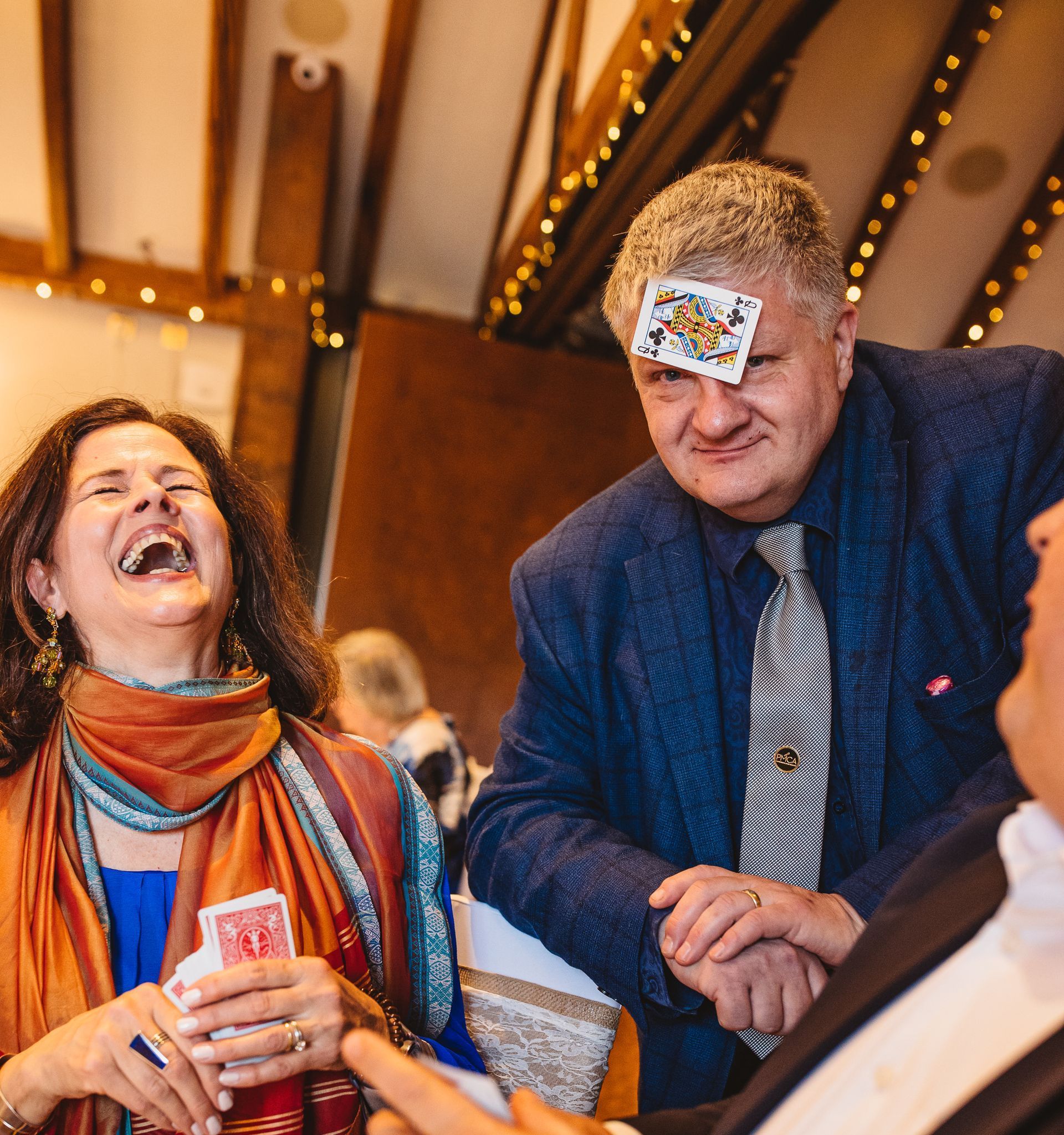 Woman laughs as a card is stuck to a man's forehead; indoor setting, possibly a party.