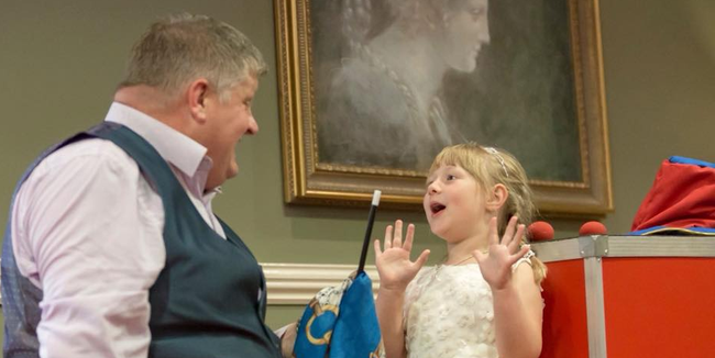A magician and a girl react with astonishment. The magician is holding a wand and blue cloth. The setting is inside.