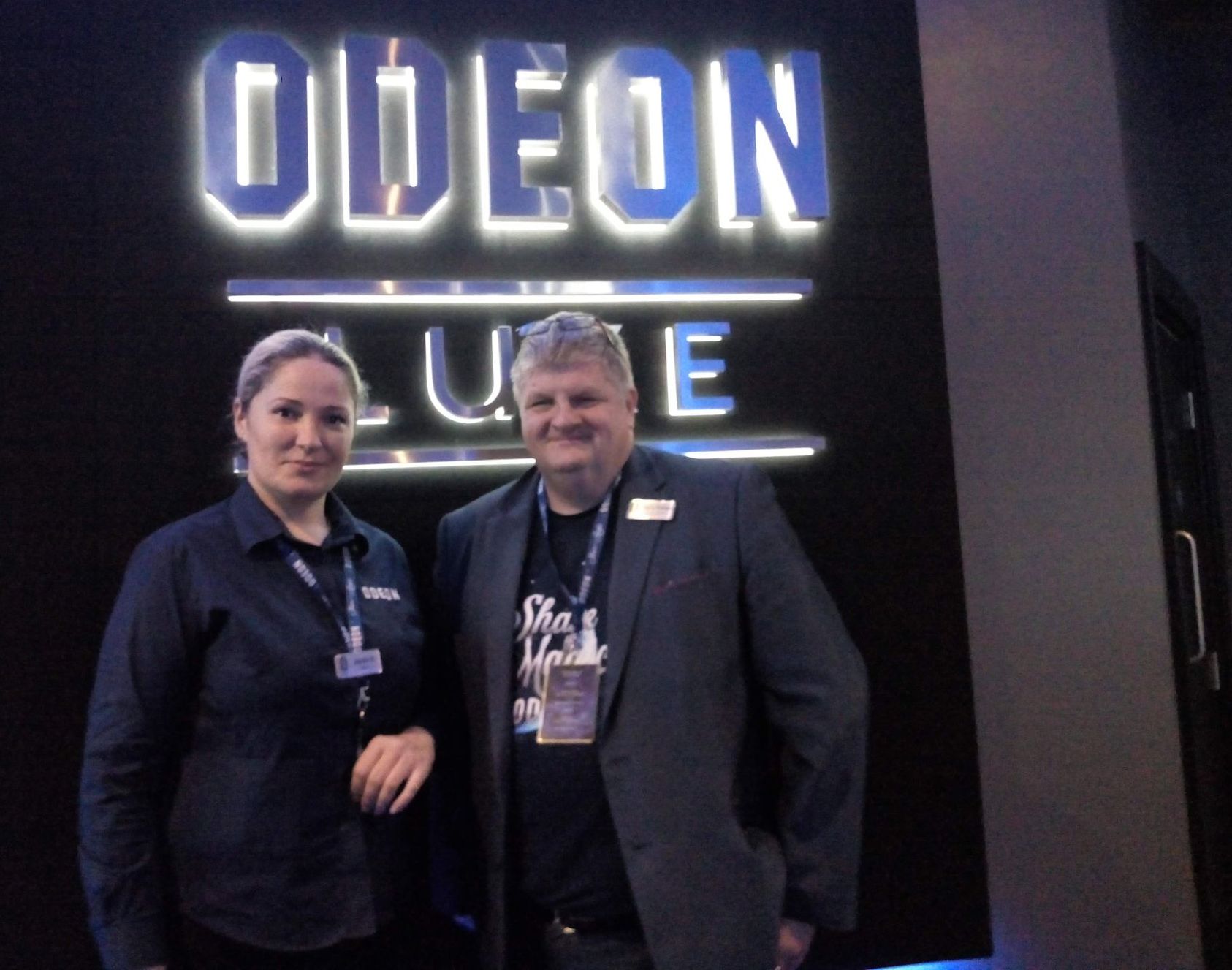 Two people stand in front of an Odeon Luxe sign. The man wears a blazer and a shirt with text. The woman wears a dark shirt.