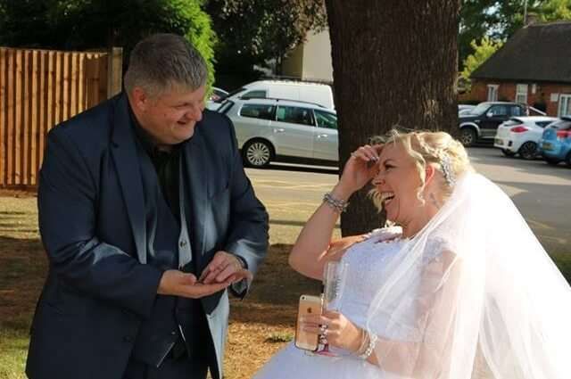 Groom and bride laughing. The groom in a suit holds out his hand, and the bride in a wedding dress touches her forehead.