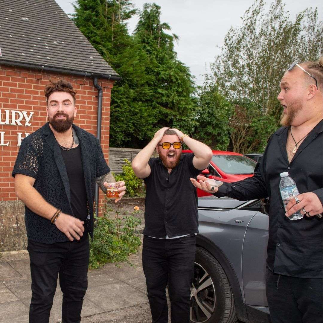 Three men outdoors: one pouring water, one with hands on head, one smiling with drink, next to a car and brick building.