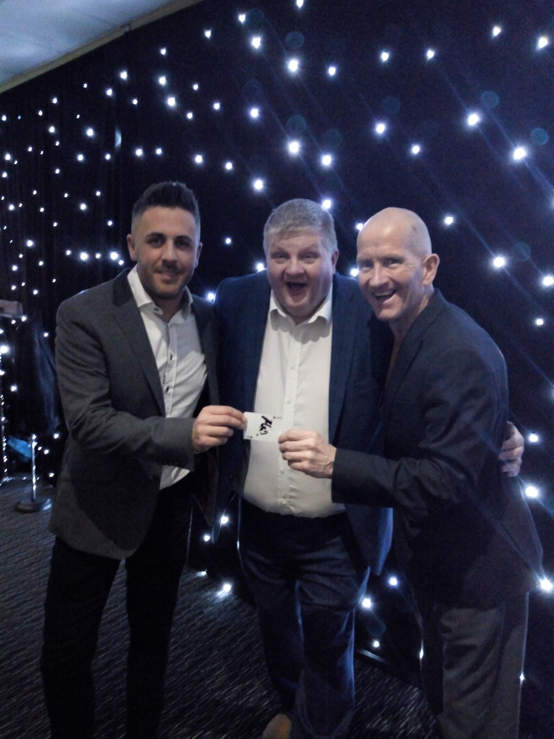 Three men smiling, holding a small card in front of a starry backdrop. One man is excited, others are looking at the card.