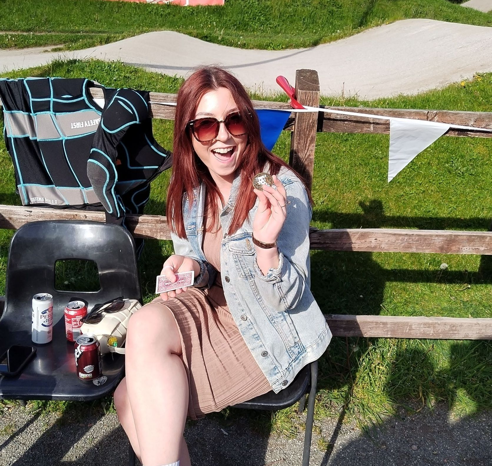 Woman in sunglasses and denim jacket, seated and smiling, holds an object in her hand outdoors near a wooden fence and grassy area.