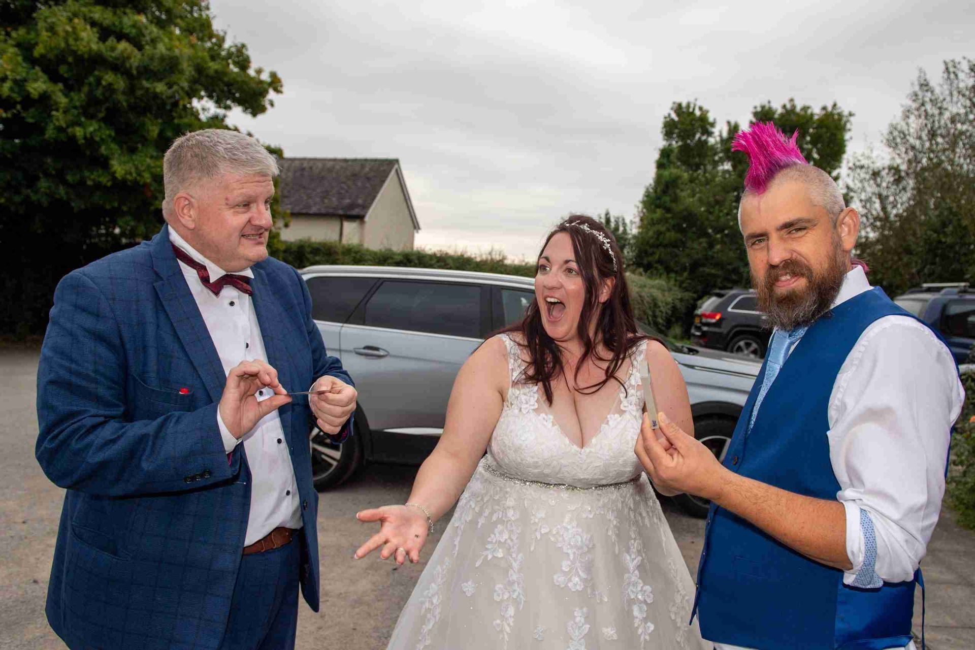A bride laughs, reacting to a magic trick by two men in formal wear outside. One man has a pink mohawk.