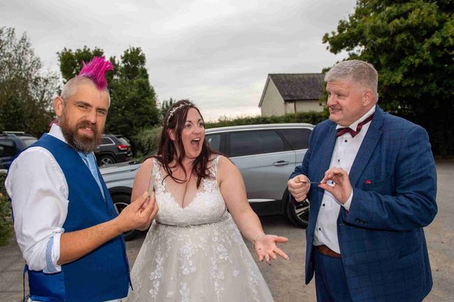 Wedding photo: A bride and two men, one with a pink mohawk, react with surprise. They are outdoors, likely at a reception.