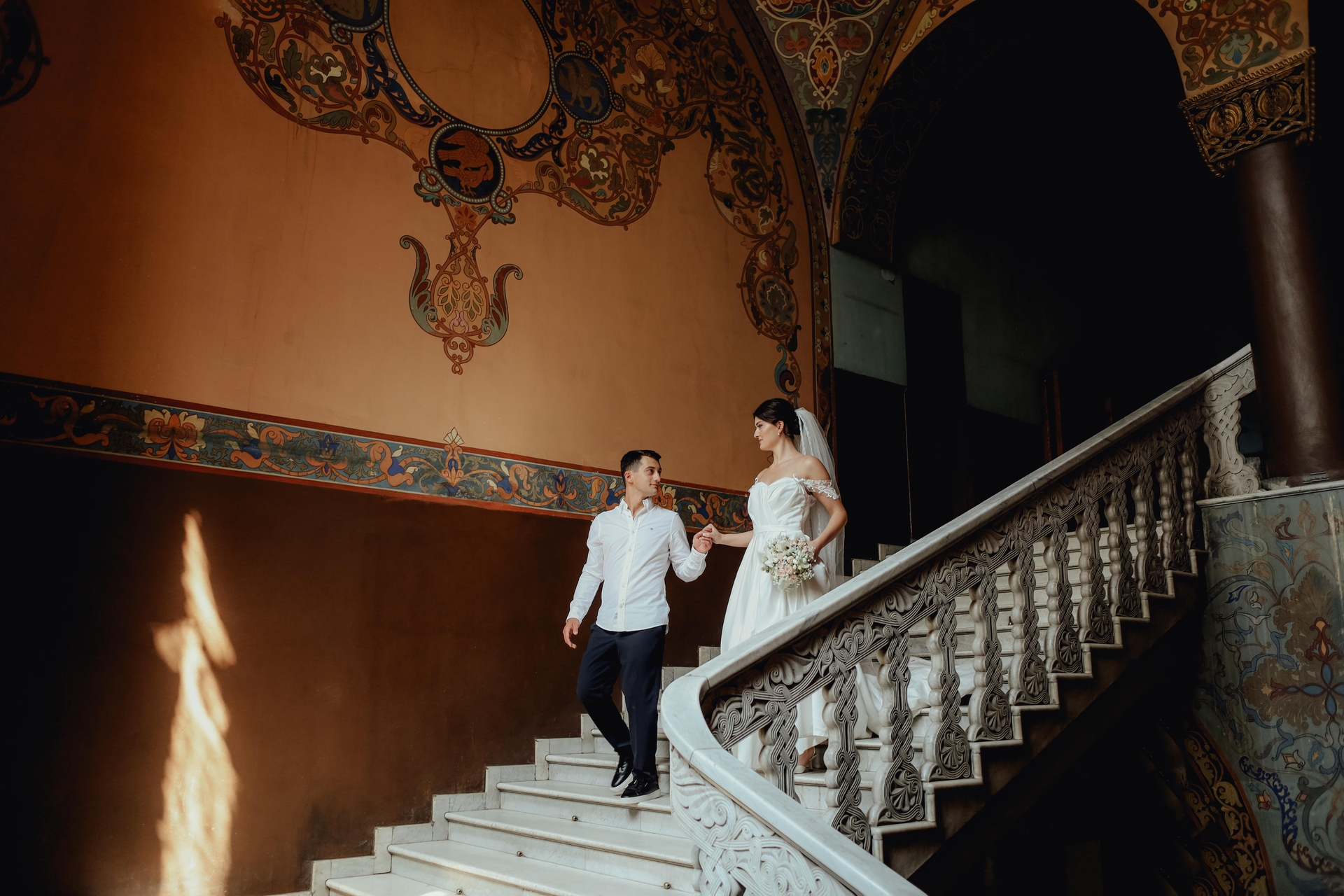 Bride and groom descend ornate staircase, holding hands, in a building with decorative wall art.