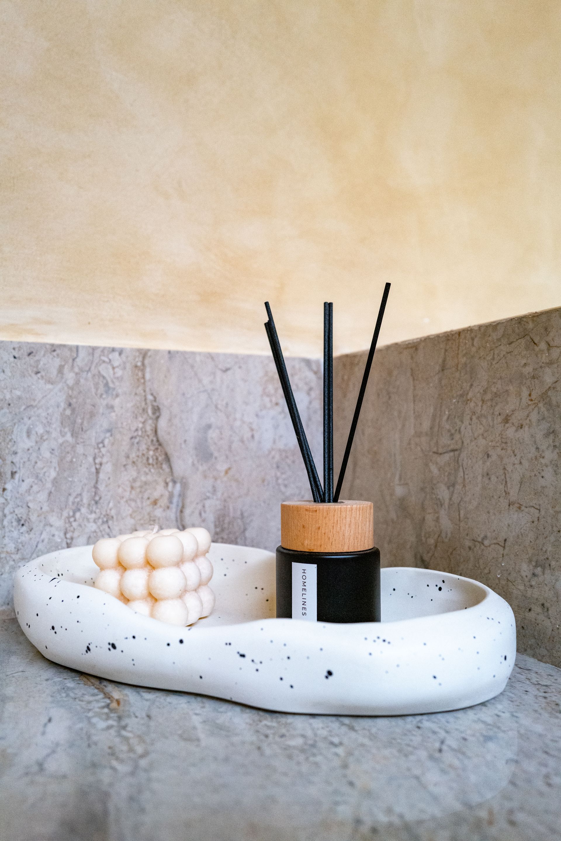 White speckled tray with candle and reed diffuser on a stone ledge, against a beige and stone background.
