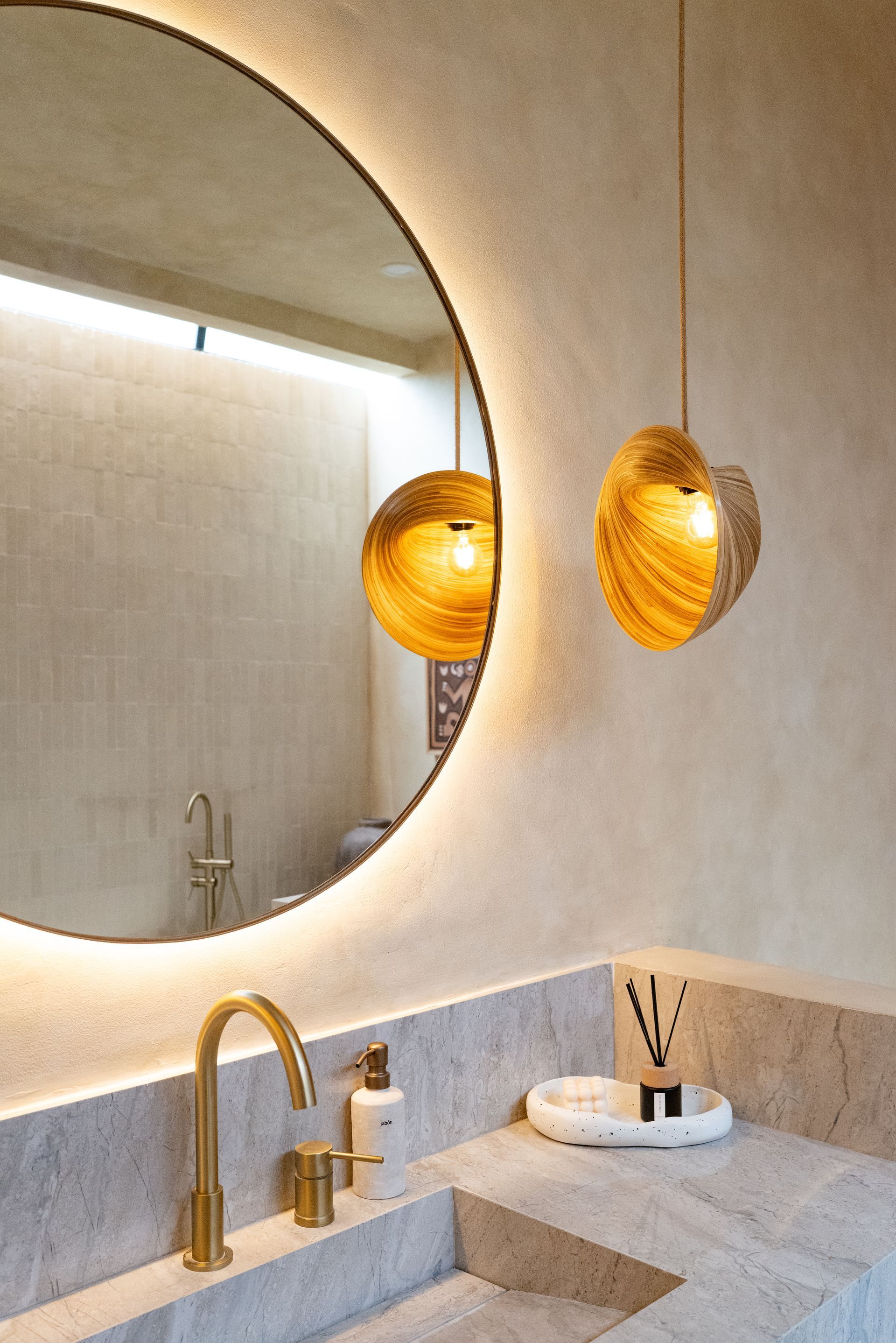 Bathroom with large round mirror, gold fixtures, and pendant lights. Beige walls, stone countertop.
