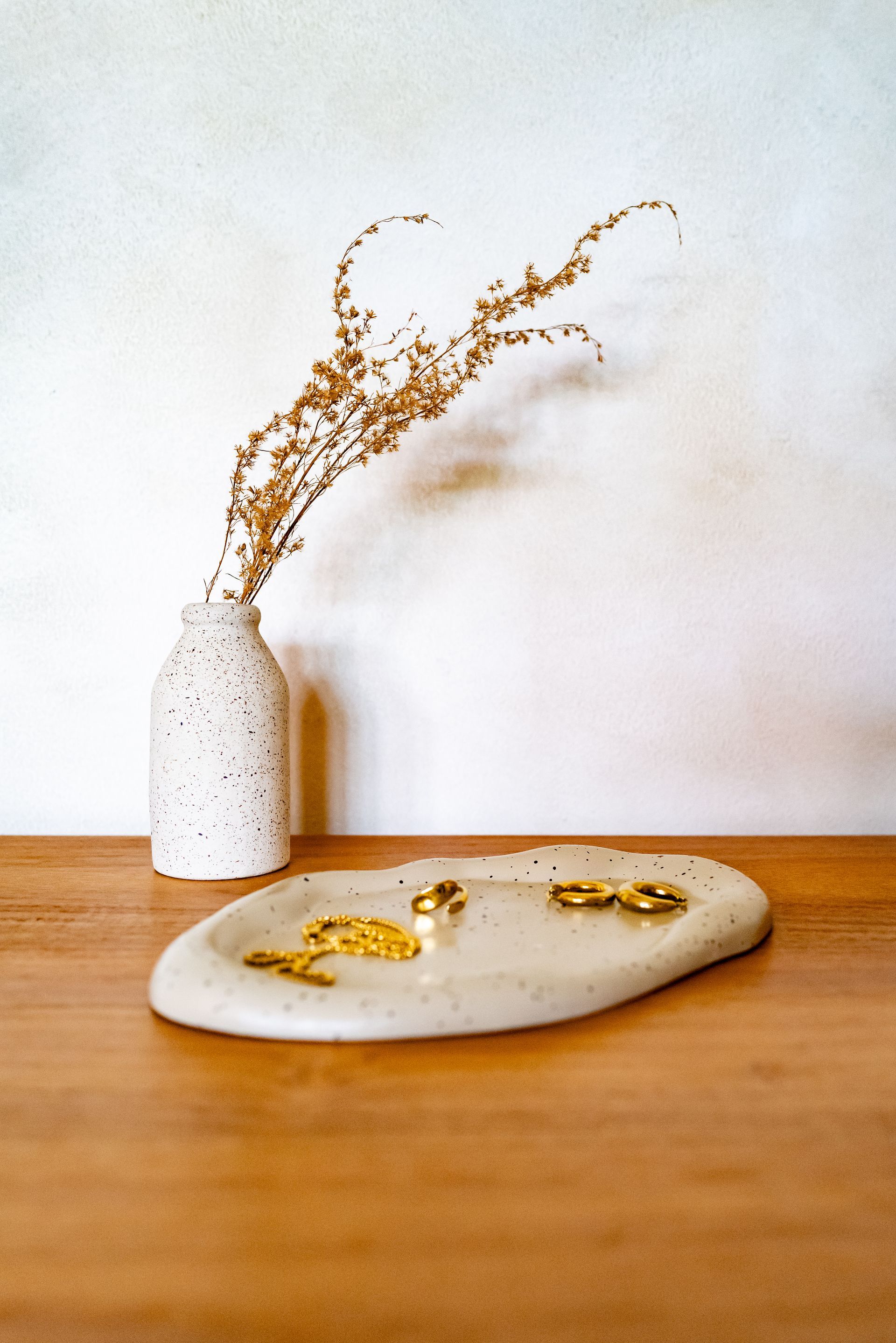 Ceramic tray with gold jewelry and speckled vase with dried flowers on a wooden table.