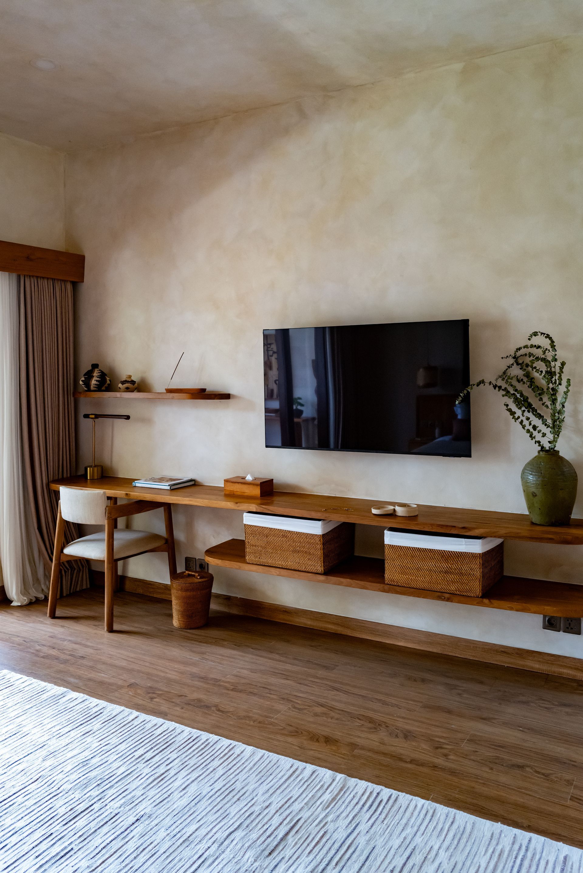 Wooden desk with TV mounted on light-colored wall, woven baskets on a shelf, and floor-to-ceiling curtain.