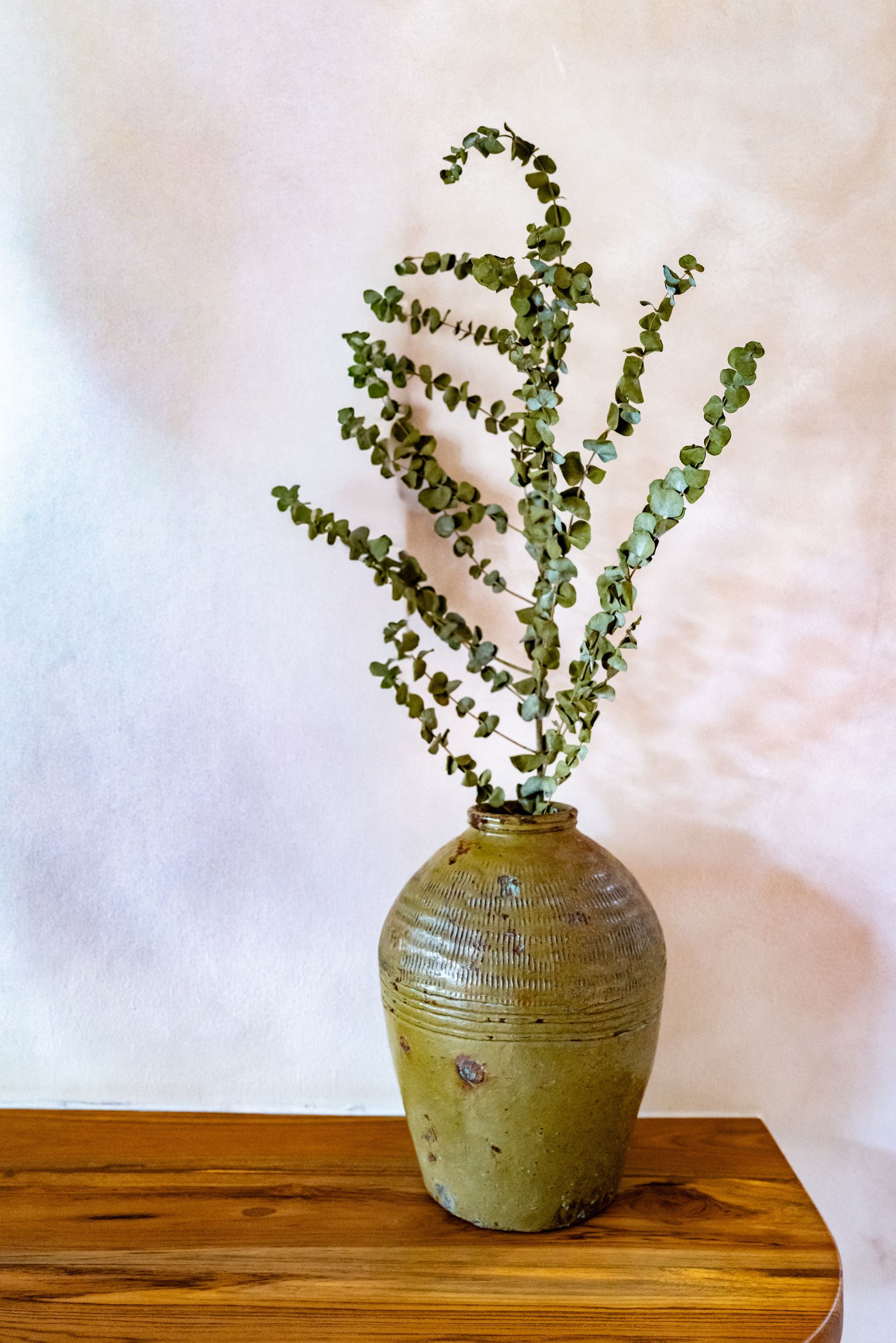 Green vase with eucalyptus on a wooden surface against a light pink wall.