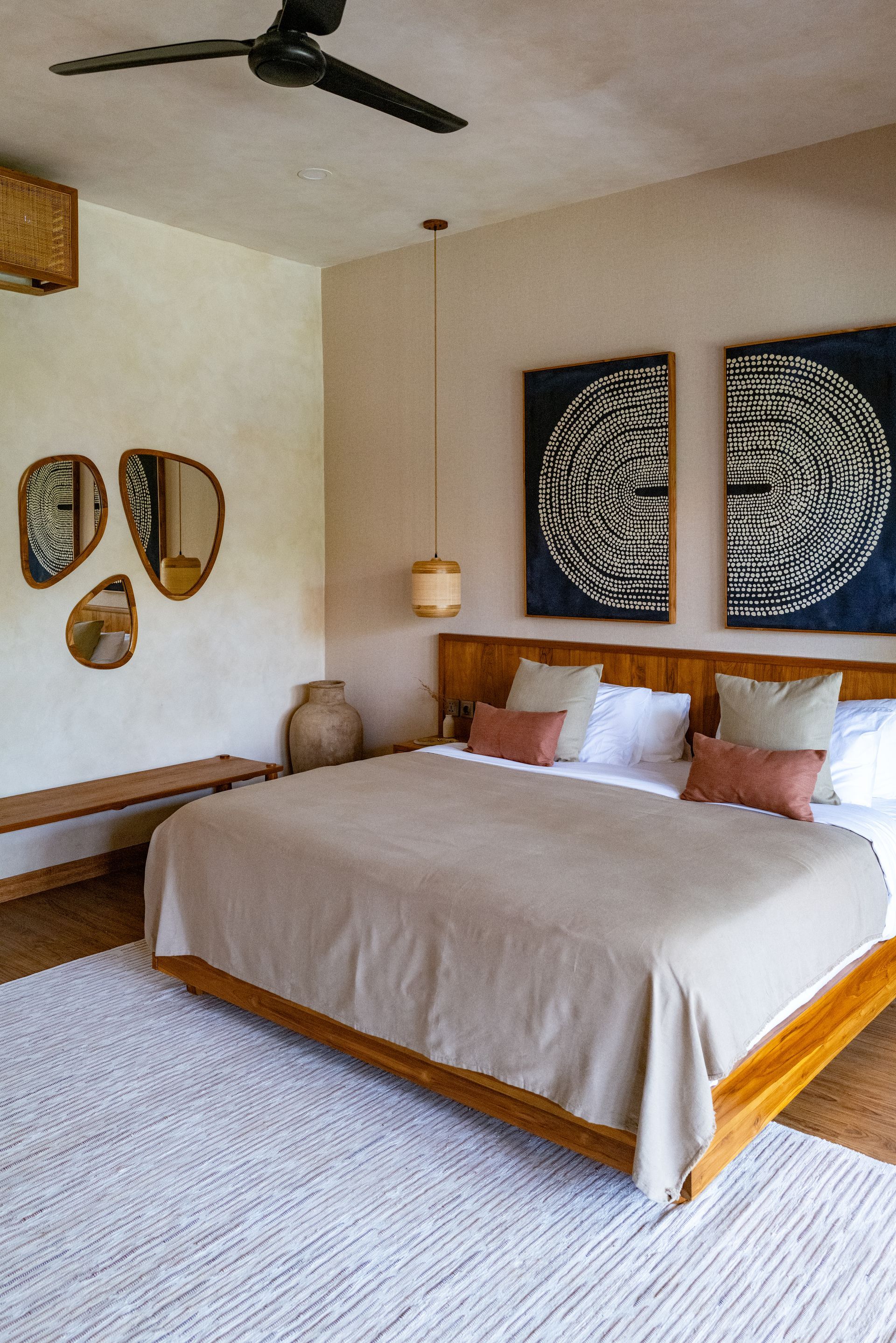 Bedroom with a wooden bed, art, mirrors, and a white rug. Neutral tones and natural light.