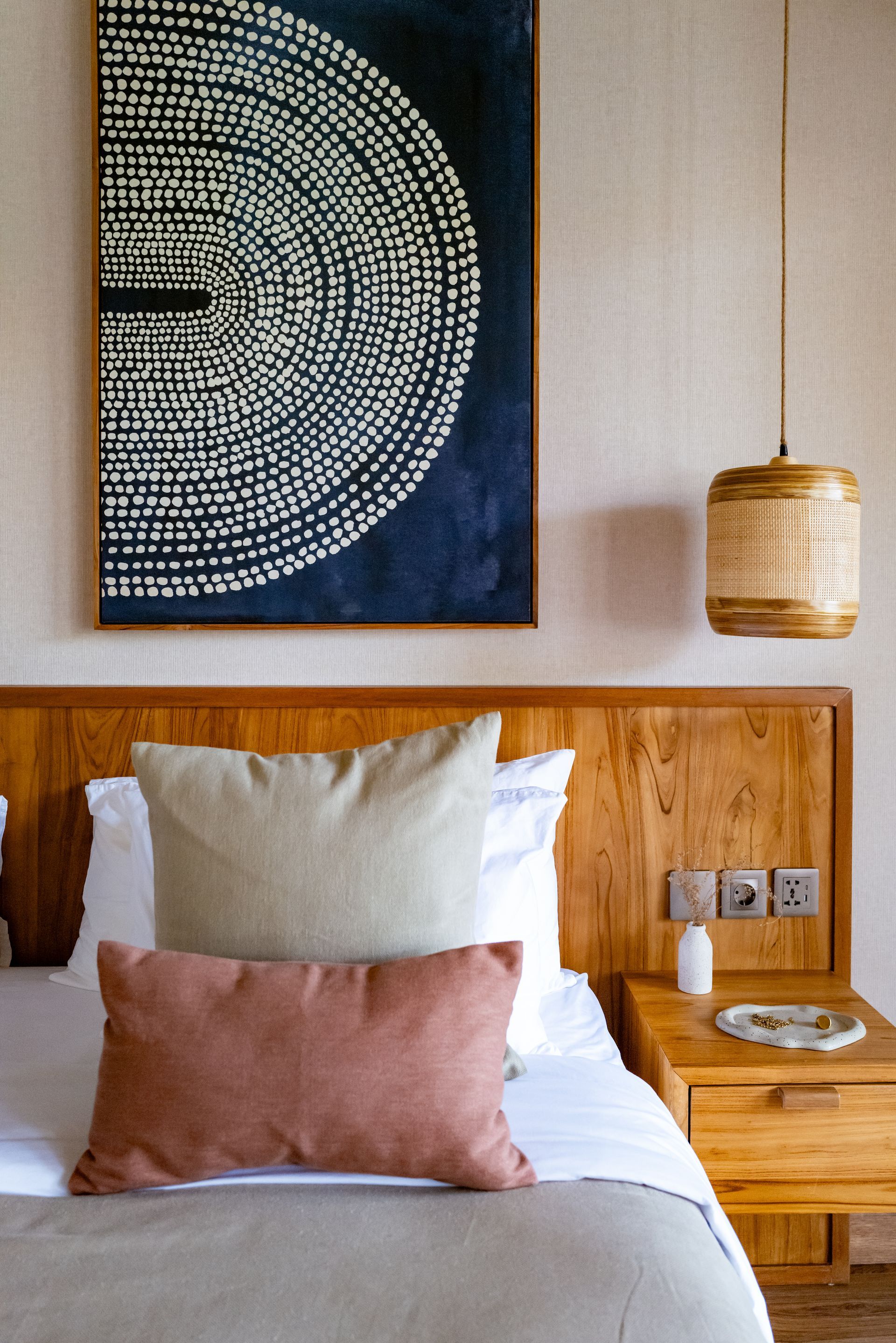 Bedroom with wooden headboard, artwork, woven pendant light, and pillows on bed.