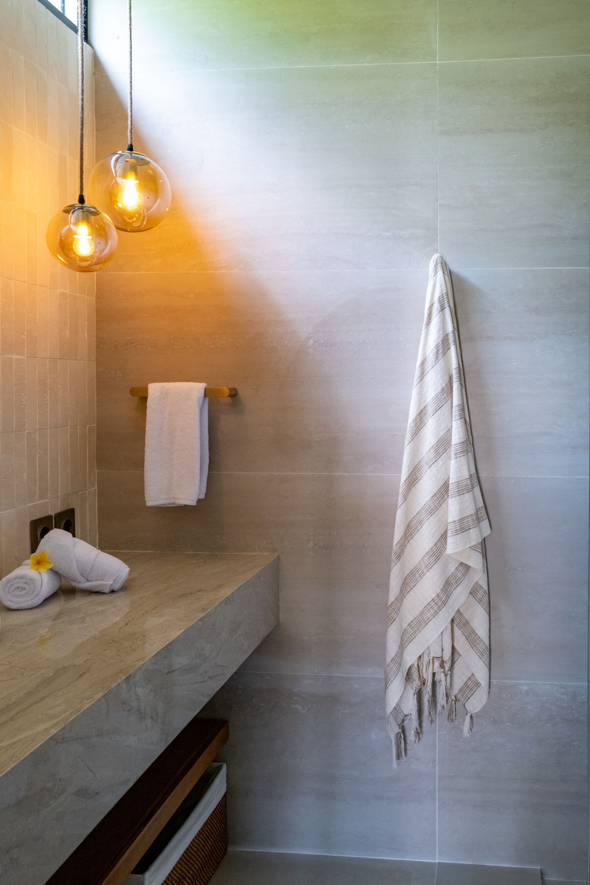 Bathroom with beige tiles, stone countertop, two pendant lights, and towels hanging.