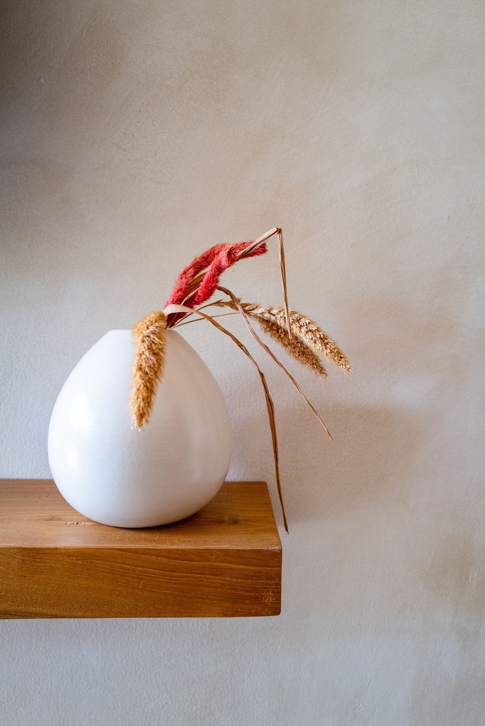 White vase with dried flowers on a wooden shelf against a beige wall.