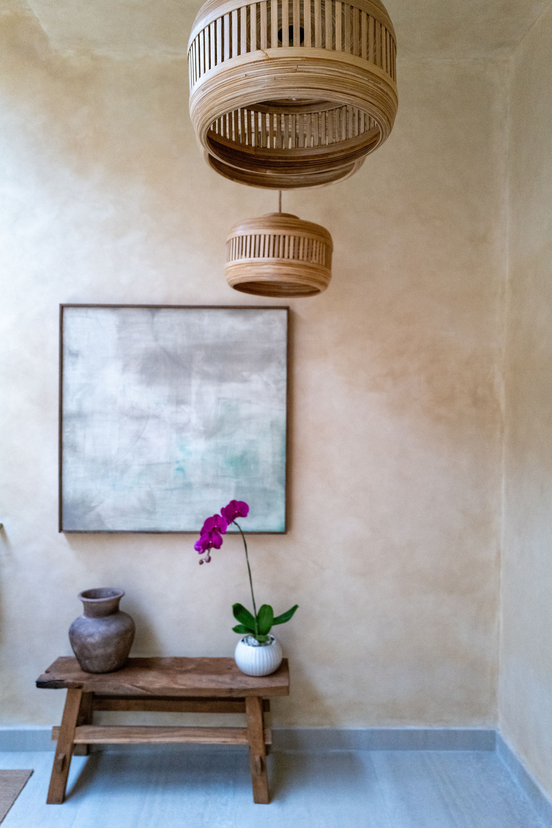 Rattan pendant lights hang above a small wooden bench with a vase, orchid, and art on a beige wall.