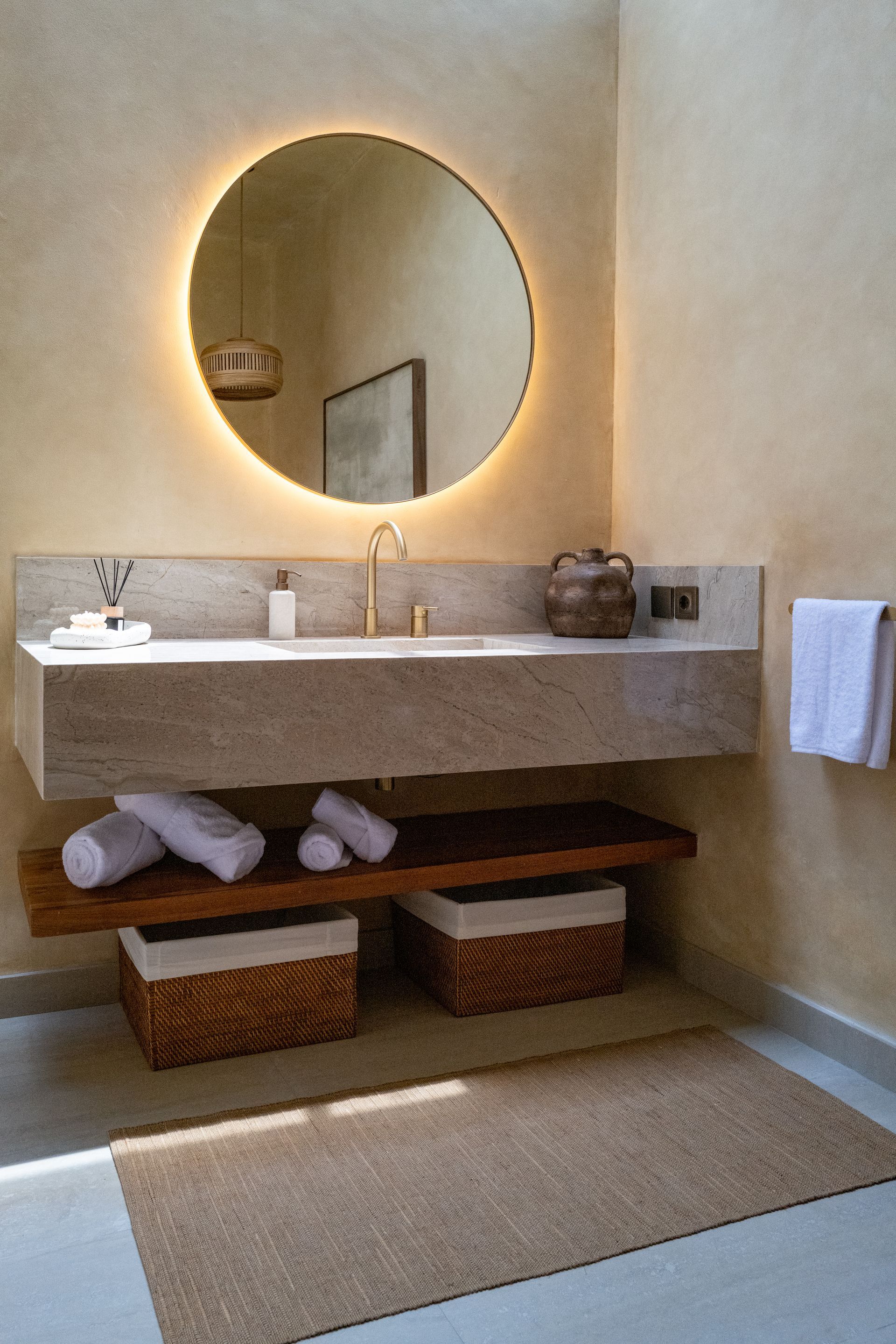 Bathroom with a floating stone sink, round mirror, wooden shelf, and natural woven baskets.