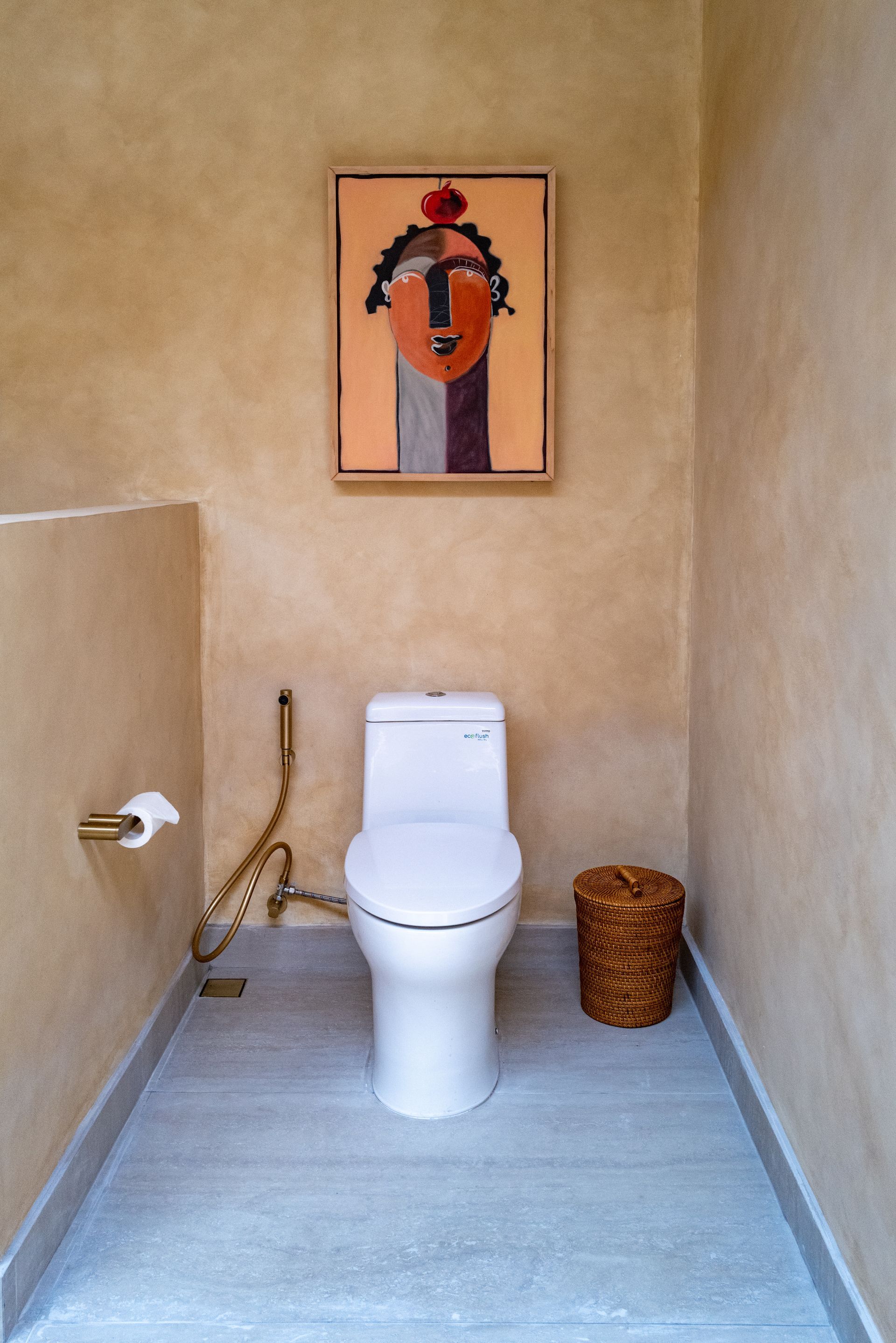 Toilet in a tan-colored bathroom. A painting hangs above the toilet. A woven basket sits nearby.