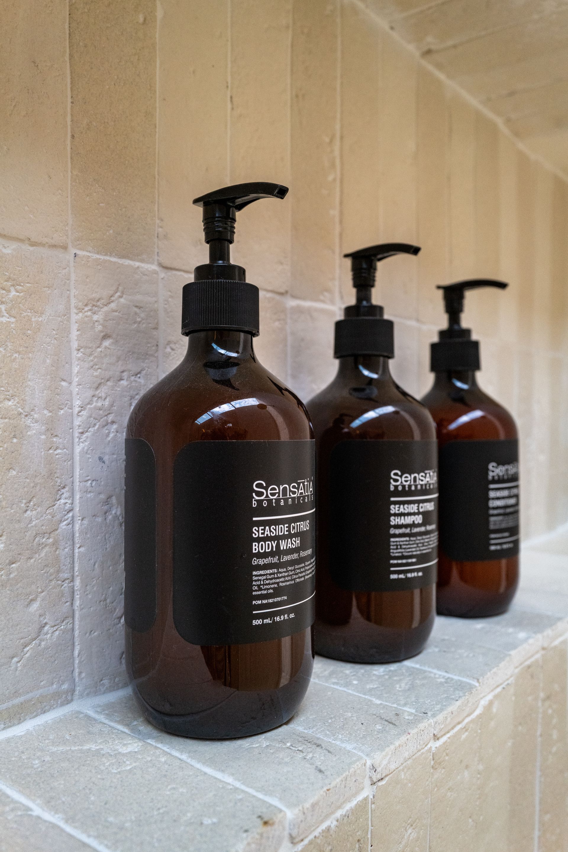Three brown pump bottles on a stone ledge in a shower, with black labels and dispensers.