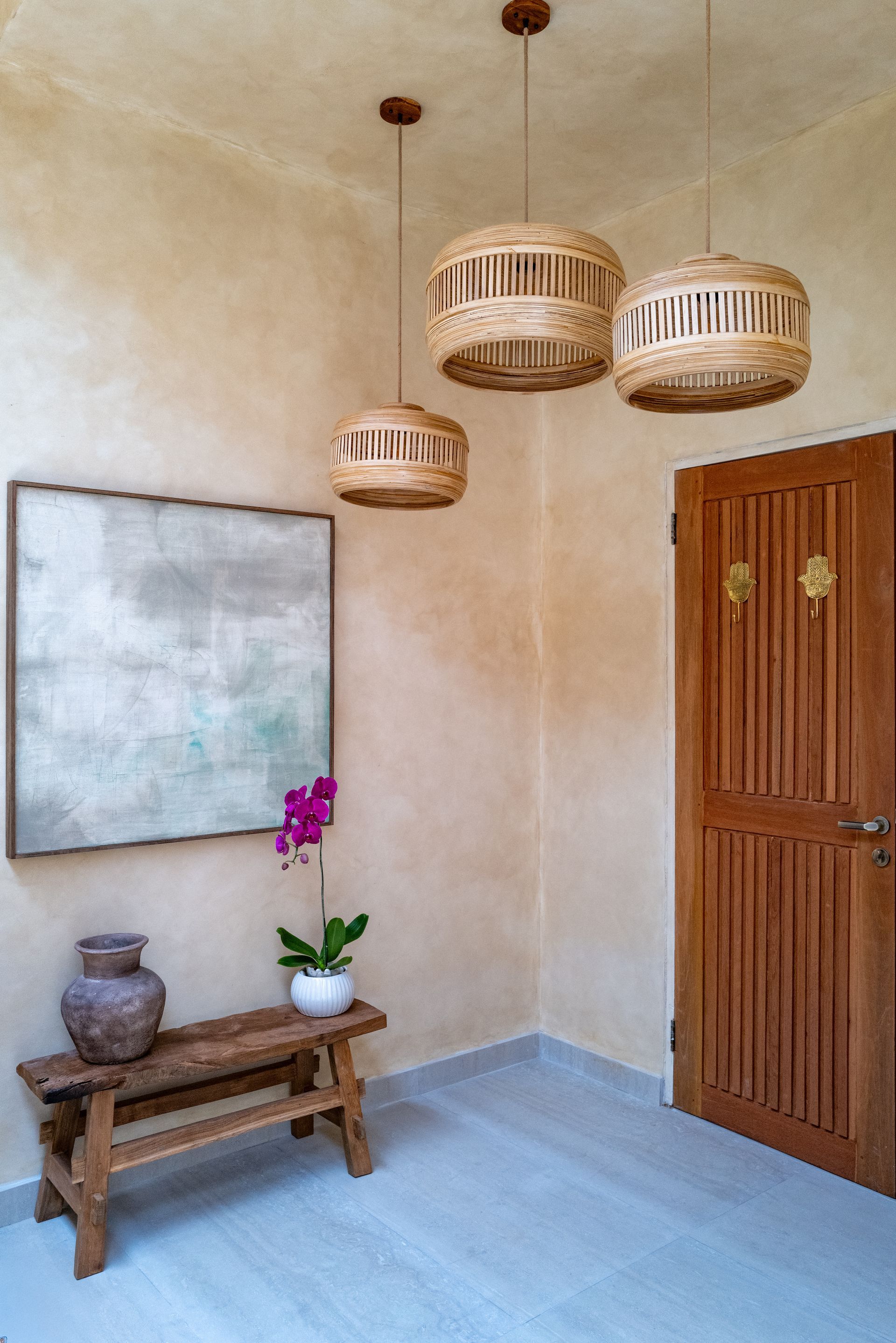 Entrance with pendant lights, art, wood door, bench, vase, and orchid. Beige walls, light blue floor.