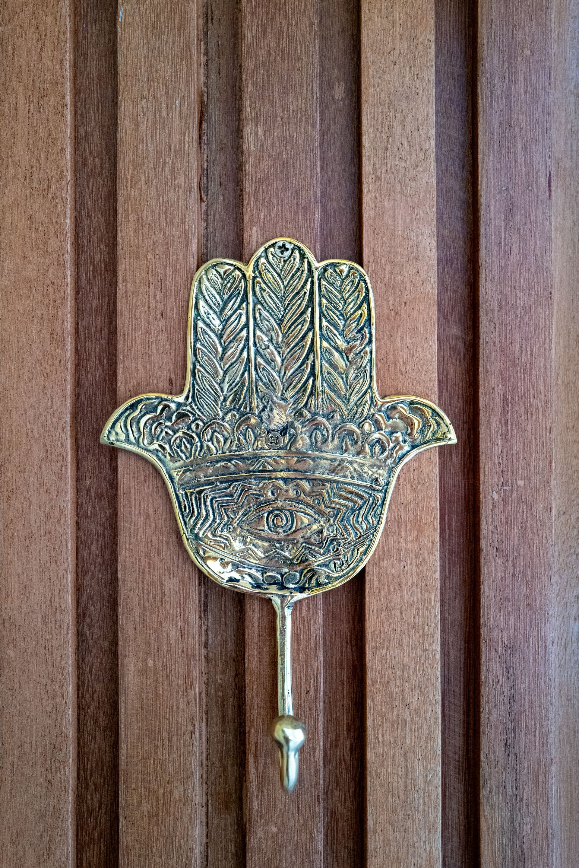 Gold Hamsa hand-shaped hook on a vertical wood-paneled door.
