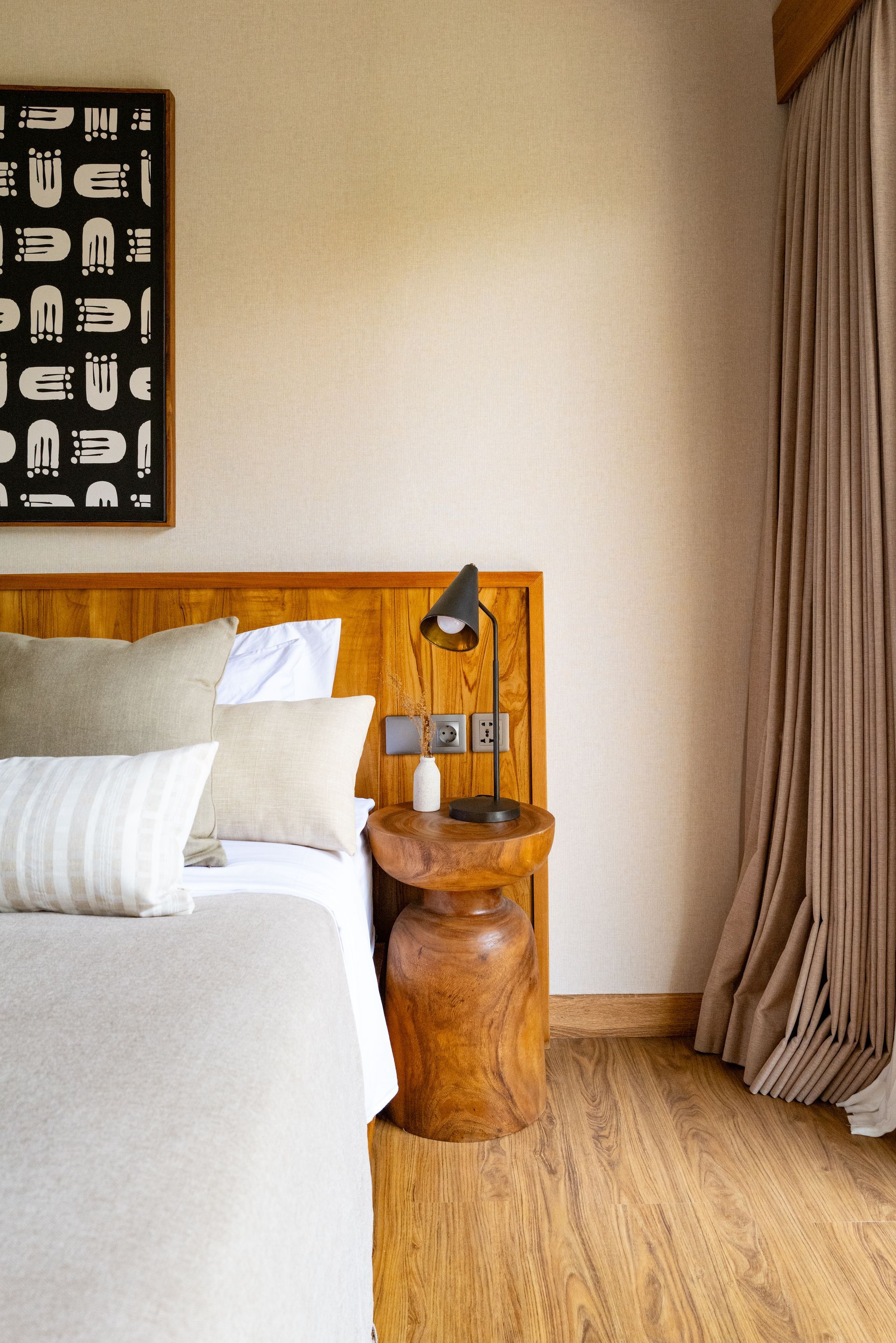 Bedroom with wooden headboard, side table, bed with pillows, art, and curtains. Earth-tone colors.