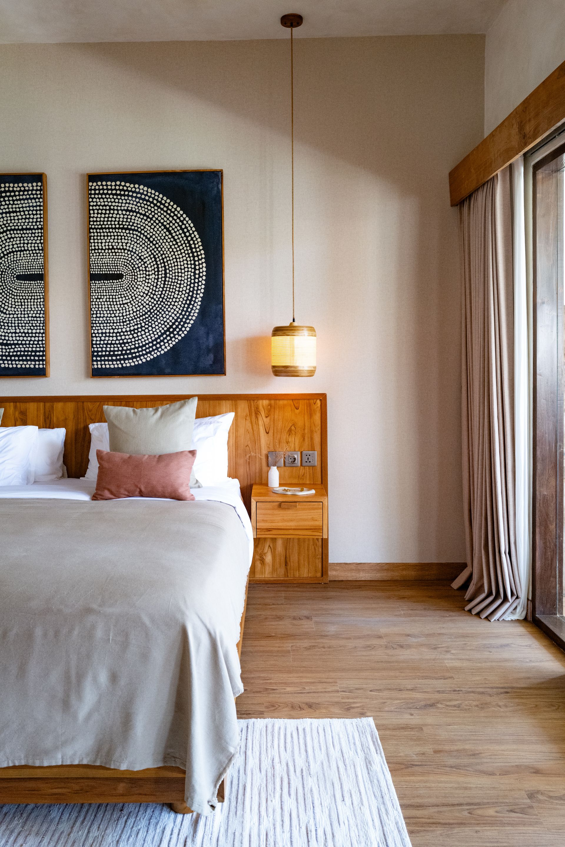 Bedroom with wooden bed, artwork, pendant light, beige walls, and wood floor.