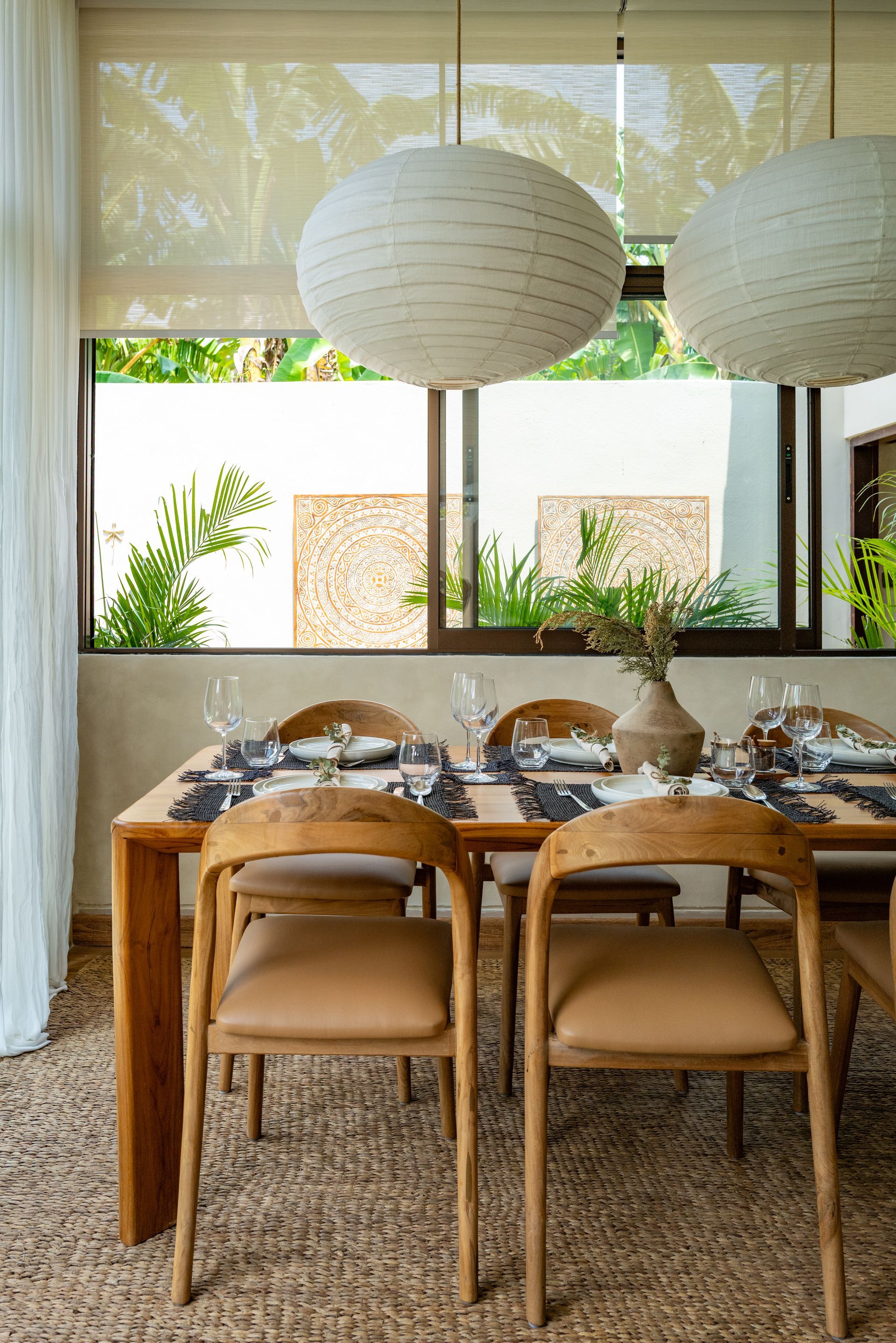Dining table with chairs beneath two paper lanterns, next to a window with greenery.