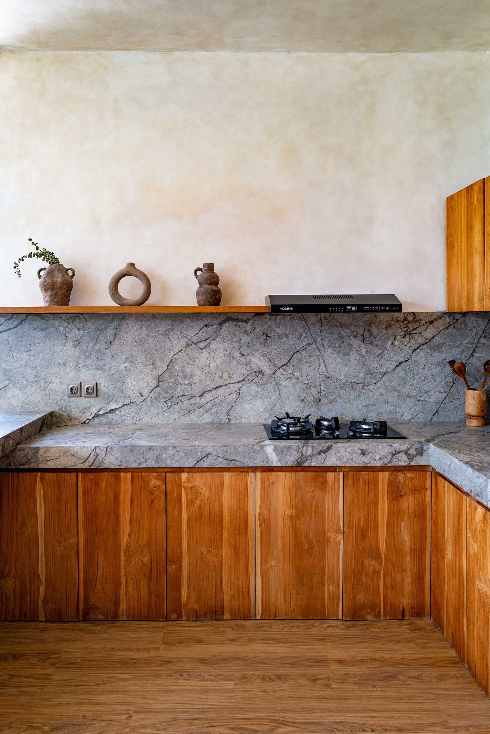 Wooden kitchen with gray countertop and backsplash, built-in cooktop, and floating shelf with pottery.