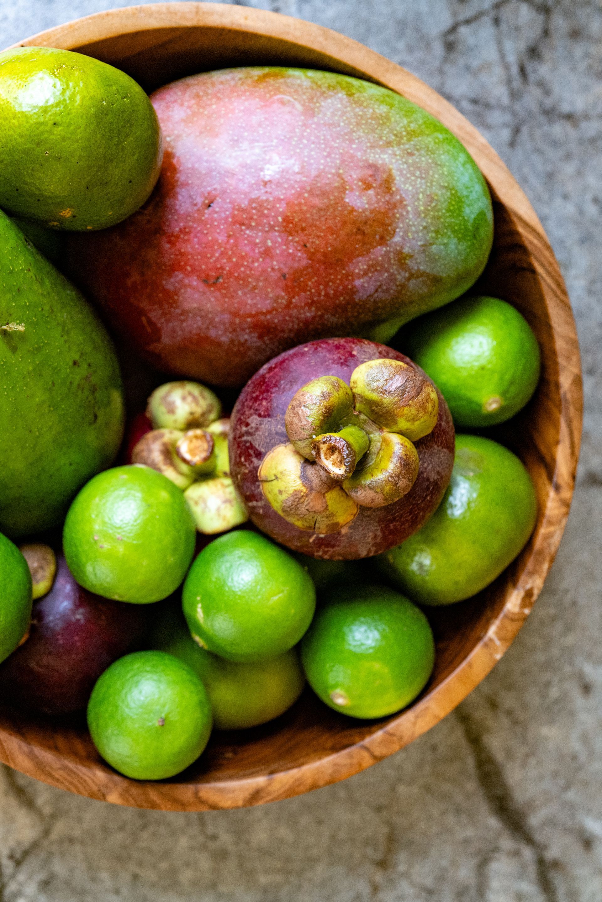 Wooden bowl overflowing with green limes, purple mangosteen, mango, and avocado.