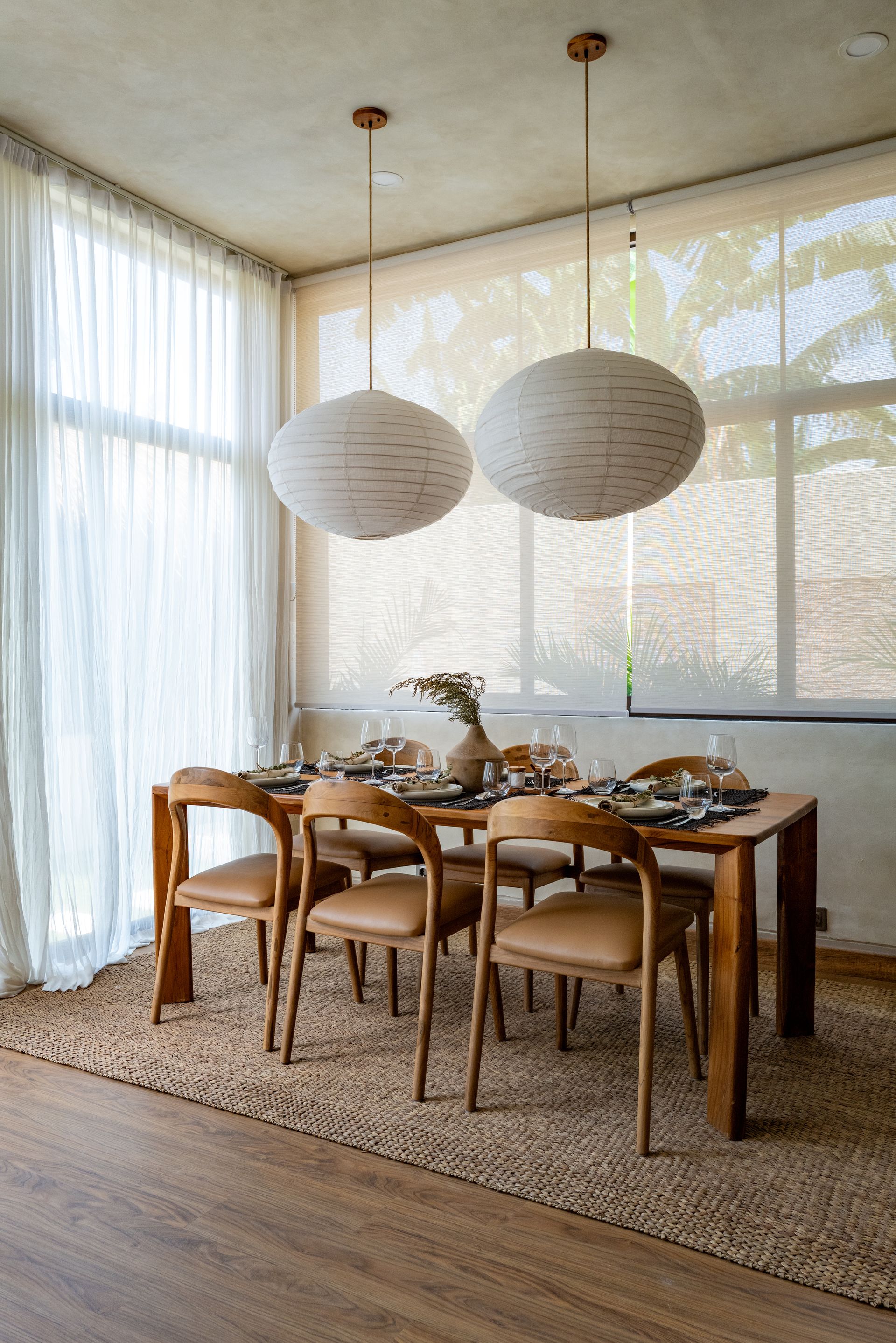 Dining room with wooden table, chairs, and two pendant lights. Sheer curtains and rug.