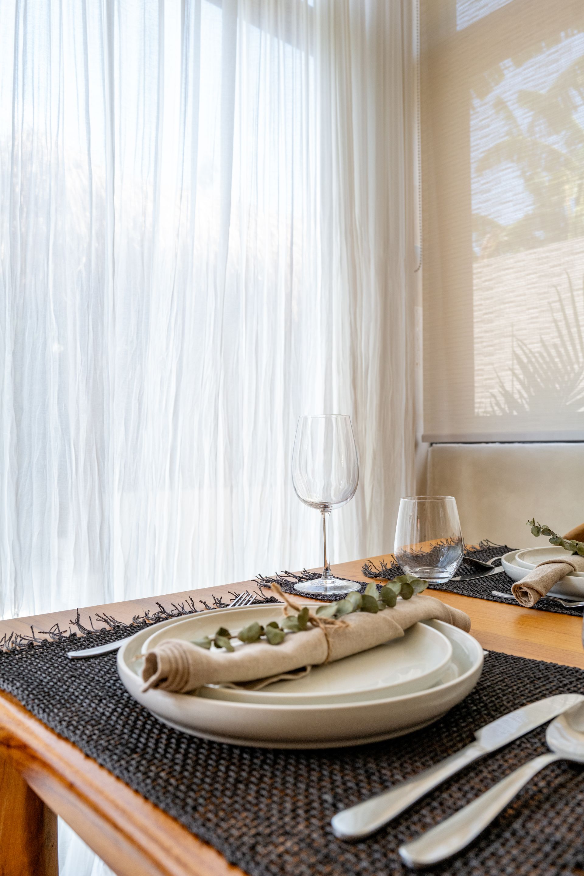 Dining table set with plates, cutlery, and a wine glass, next to a window with white curtains.