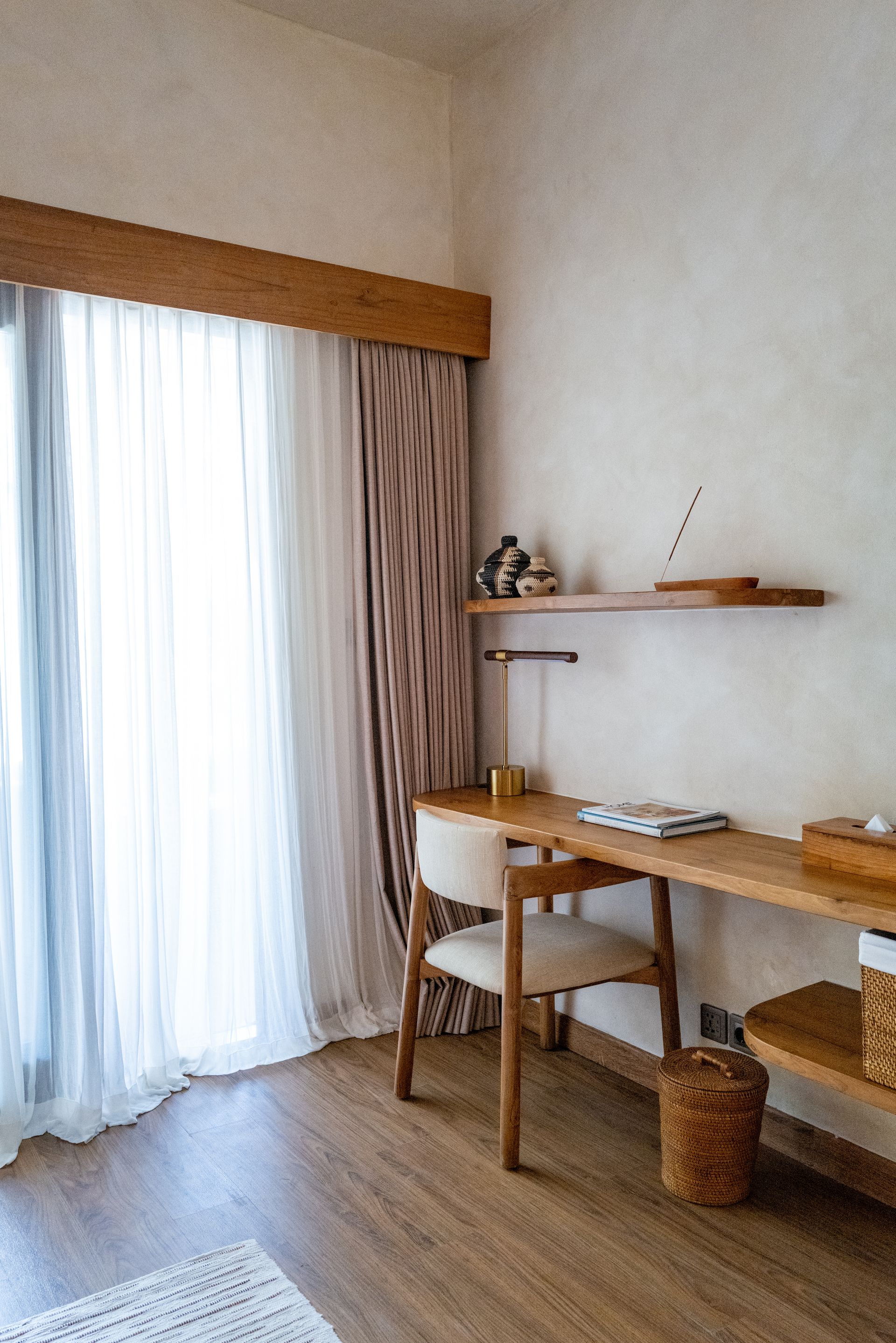 Desk and chair in front of a window with sheer curtains. Wooden shelves and a woven basket complete the minimalist room.
