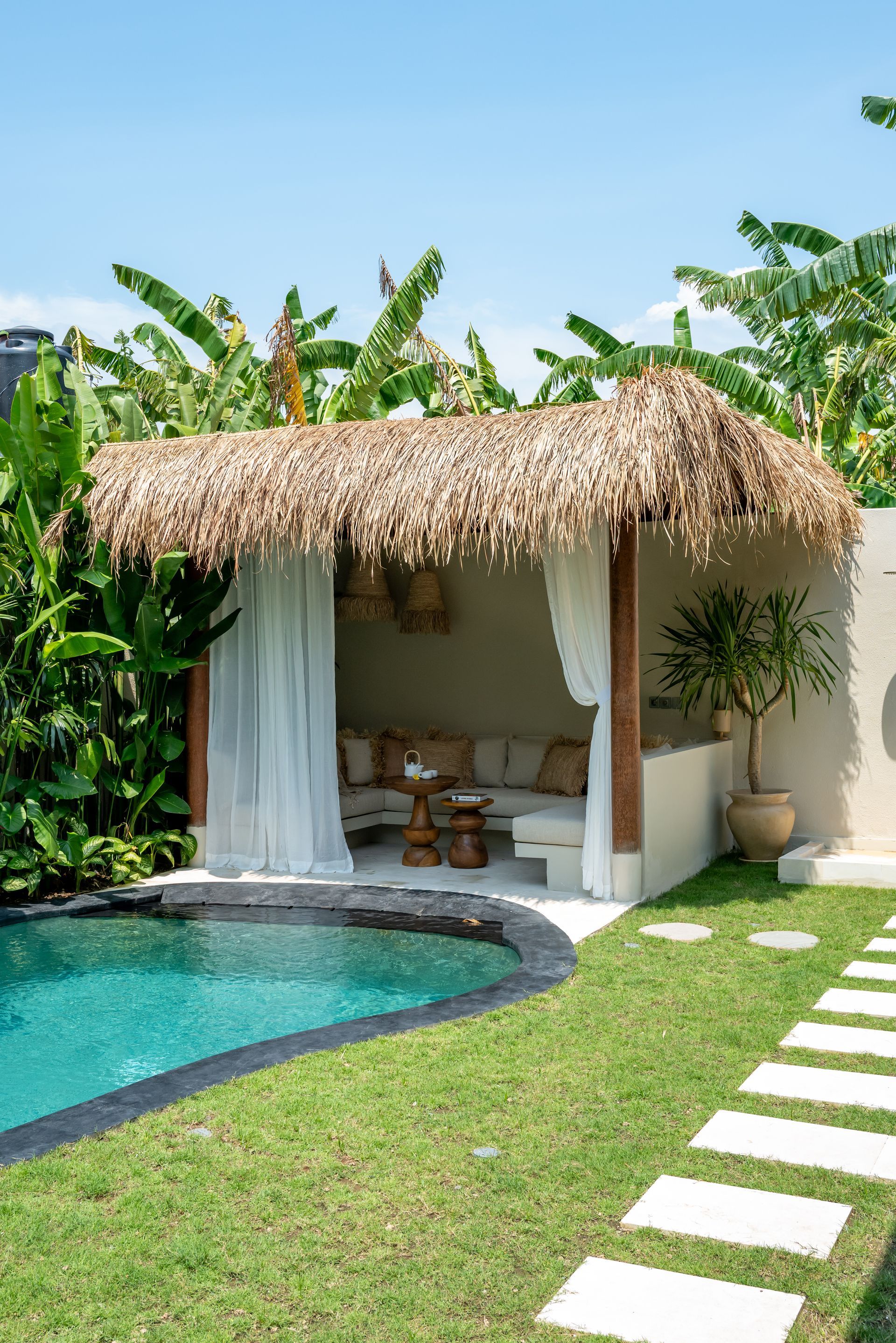 Poolside cabana with thatched roof, curtains, and cushions; lush green foliage and blue sky.