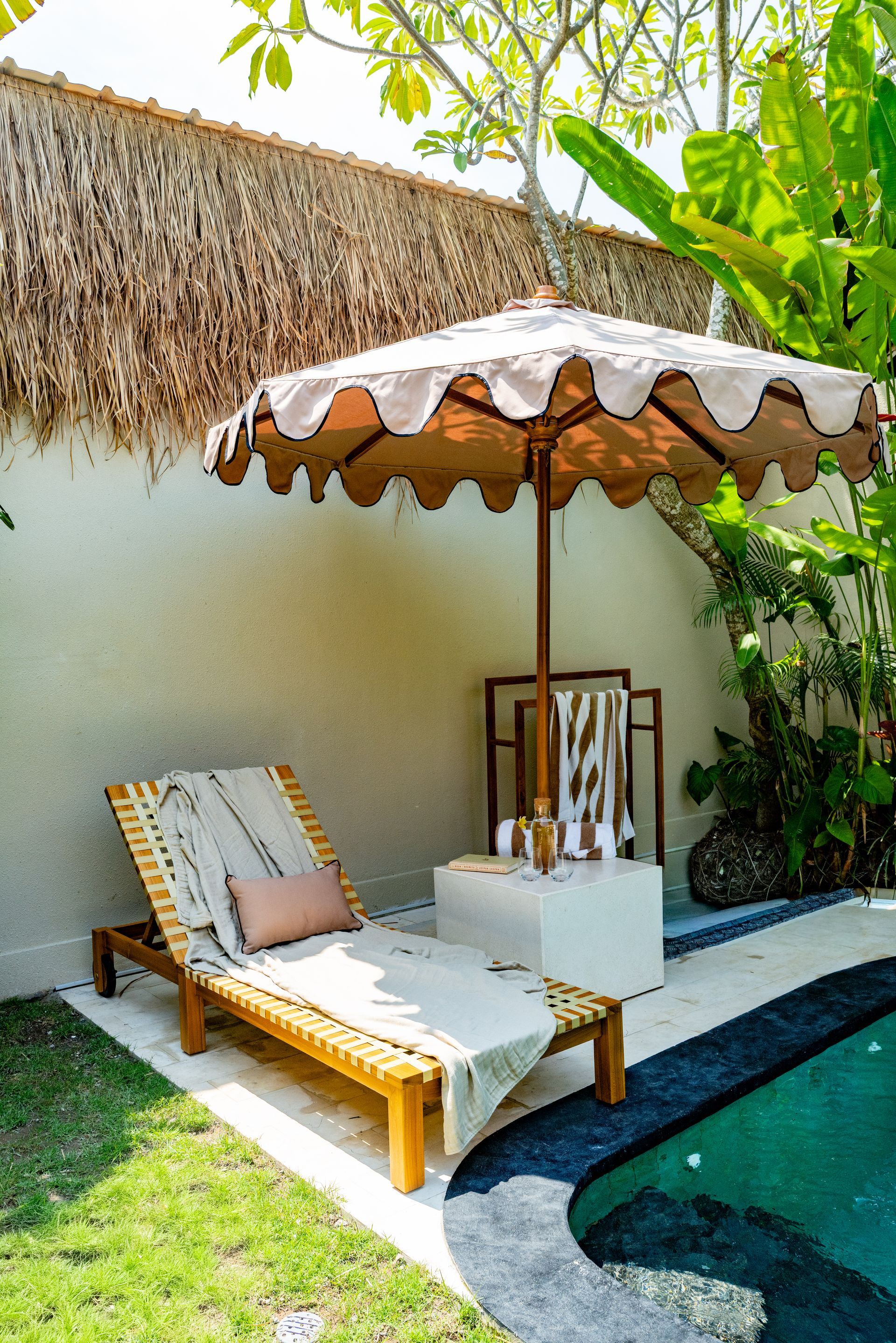 Lounge chair and umbrella by a pool. Tan chair, white umbrella, cream towels, and green foliage.