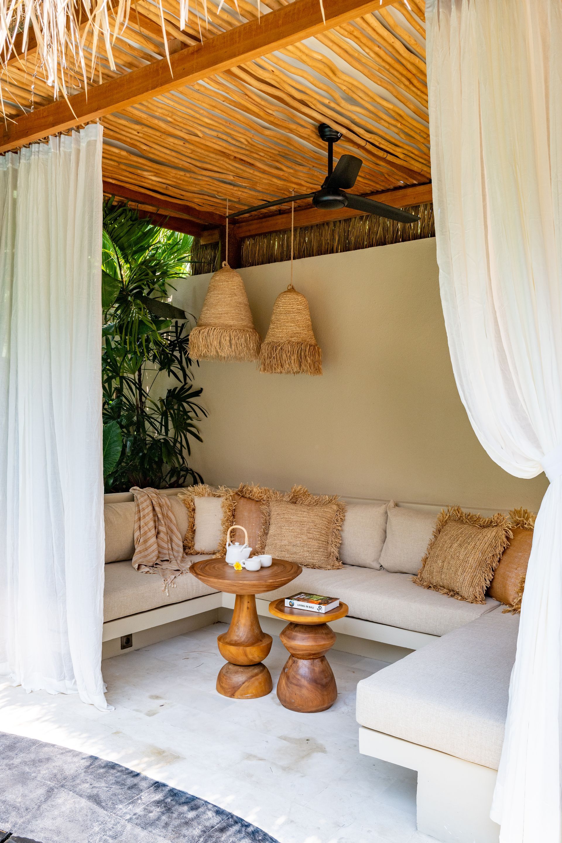 Outdoor lounge with beige cushions, woven accents, straw roof, and sheer curtains.