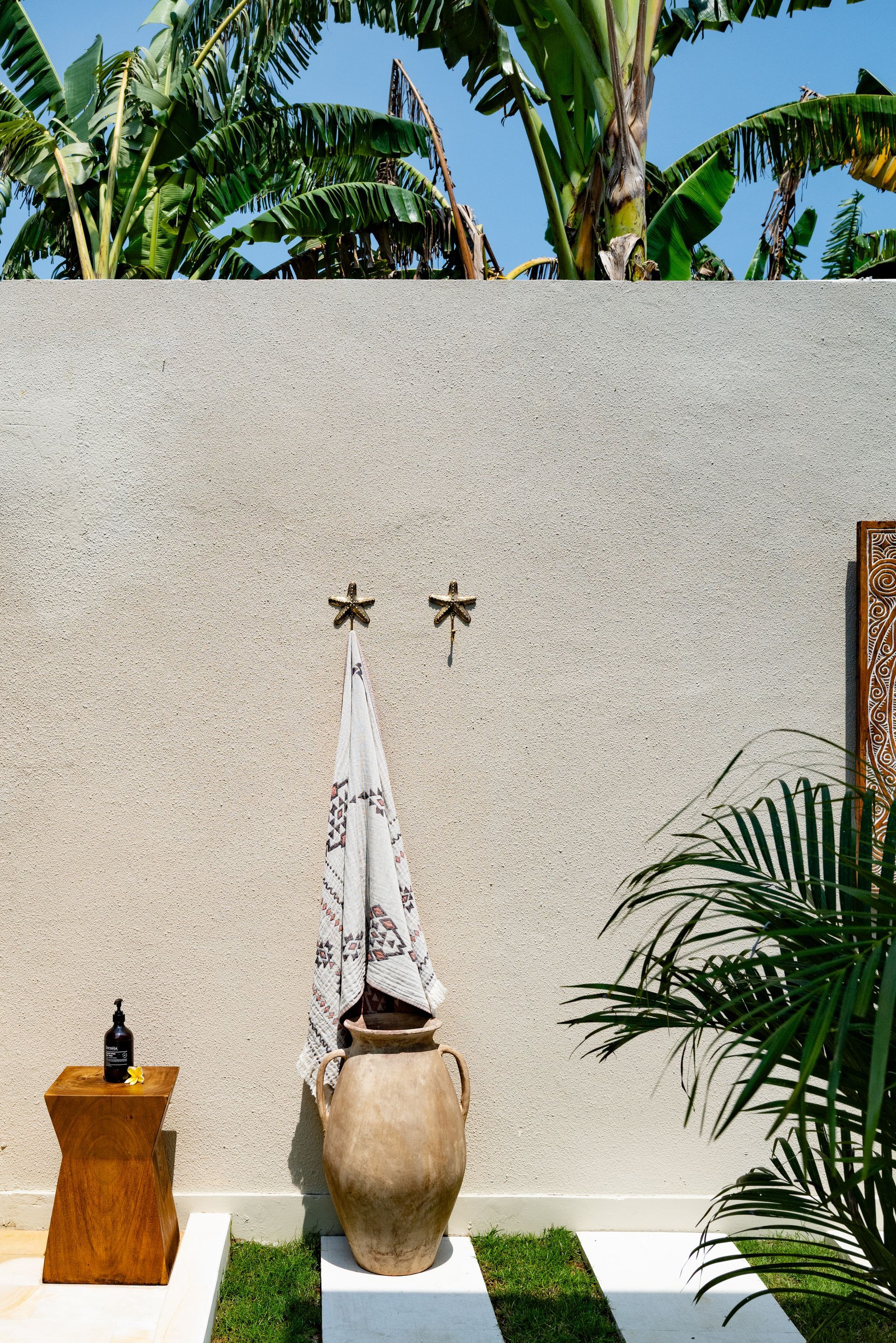 Outdoor shower with beige stone wall, towel, and large clay pot. Wooden stool with dark bottle, greenery and banana trees.