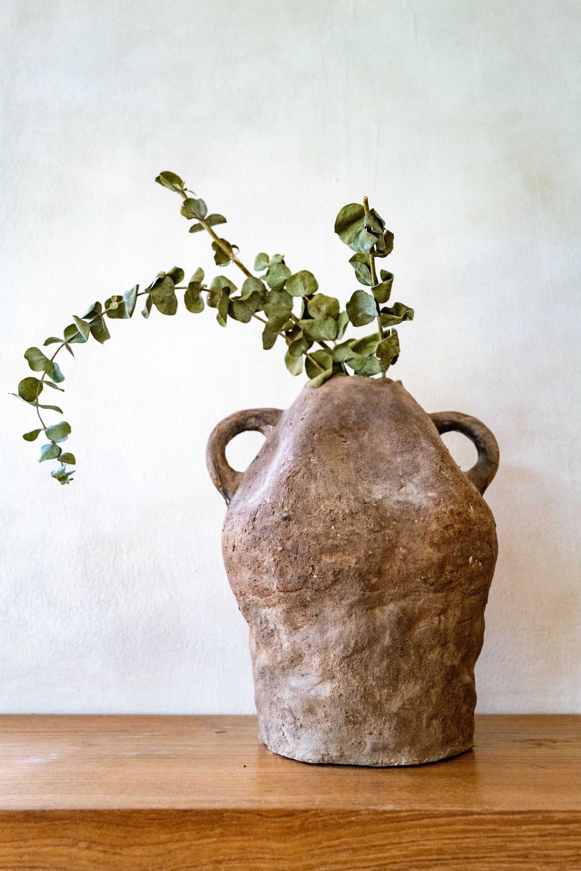 Rough, brown ceramic vase with eucalyptus branches on a wooden surface against a neutral wall.