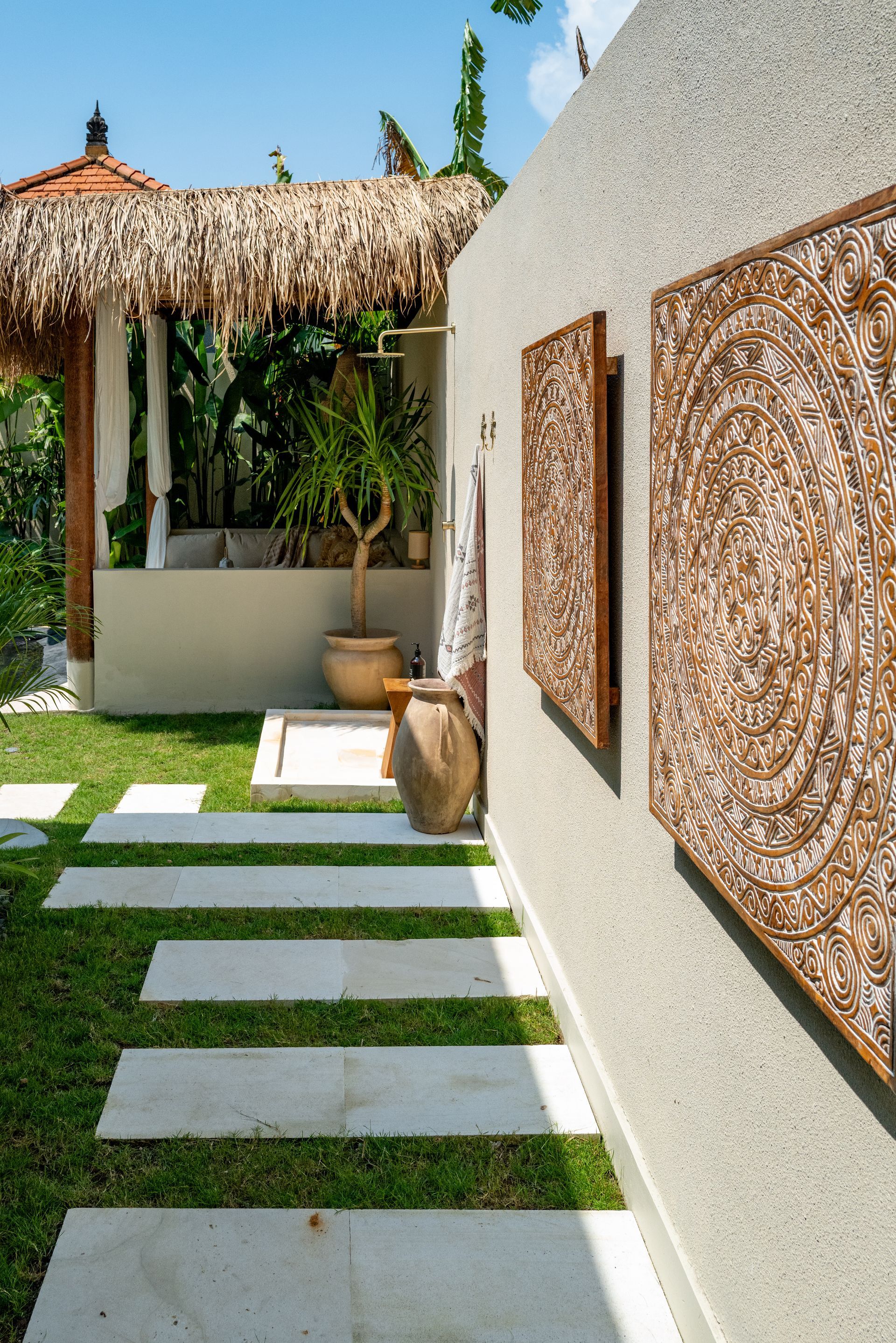 Pathway of stone slabs leads to a gazebo with thatched roof, next to a textured white wall with decorative panels.