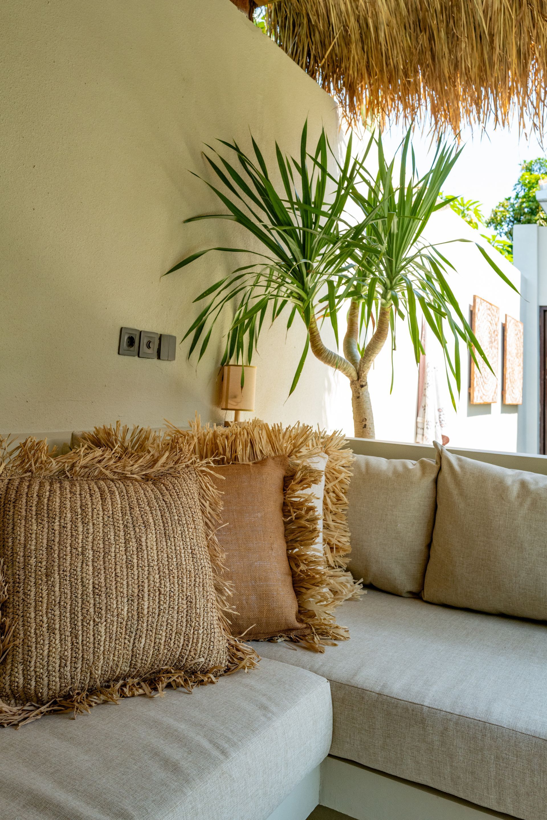 Cozy outdoor seating area with fringed pillows, light-colored cushions, and a small palm tree.