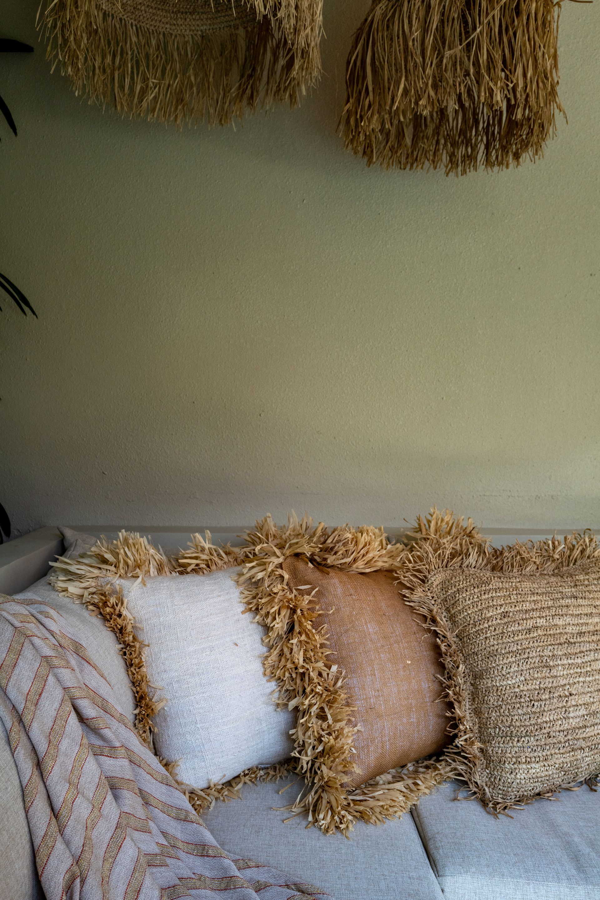 Couch with textured pillows and blanket. Beige, gold, and white tones. Natural light. Decorative straw hanging above.