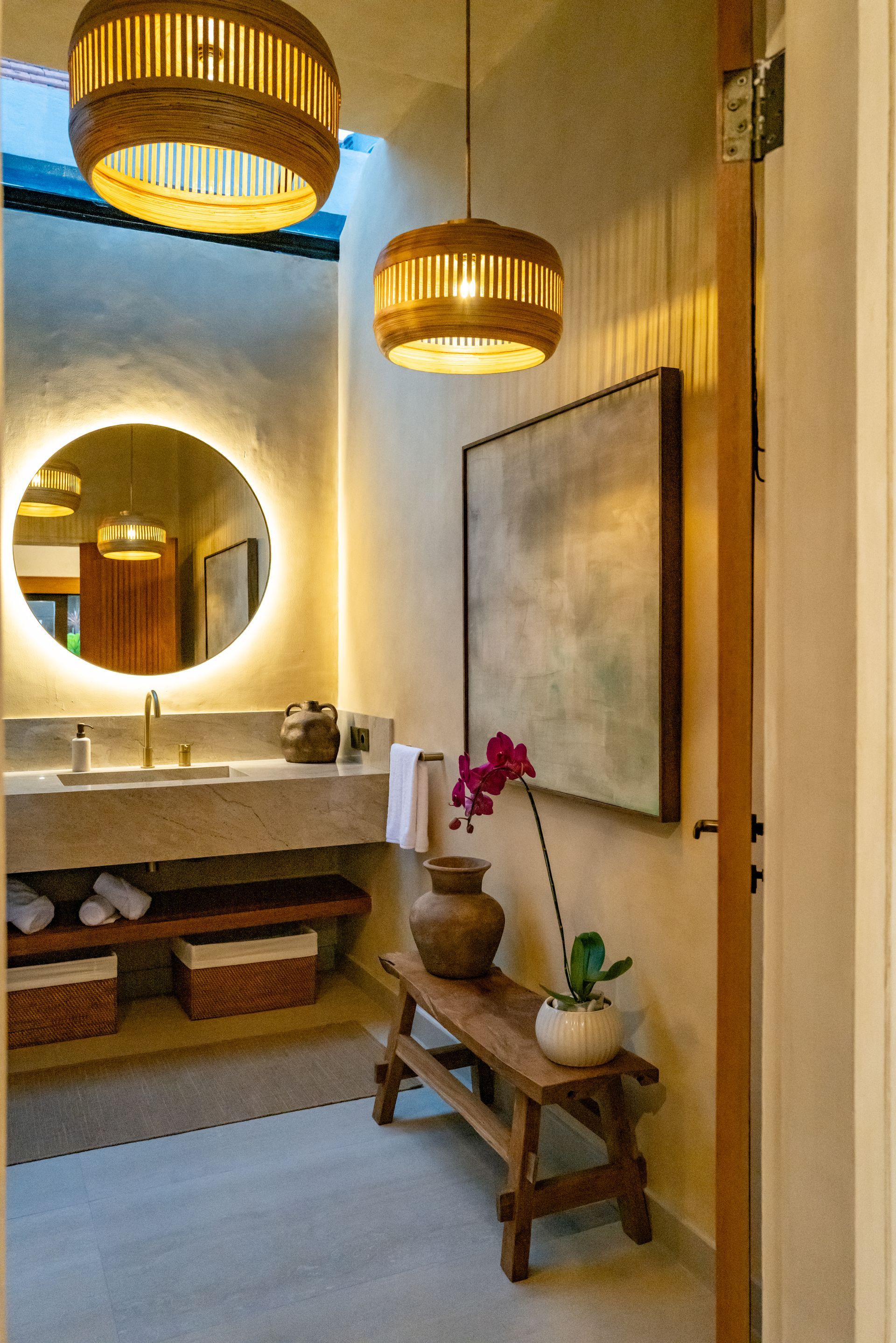 Bathroom with a round lit mirror, stone sink, wooden bench, and woven pendant lights.