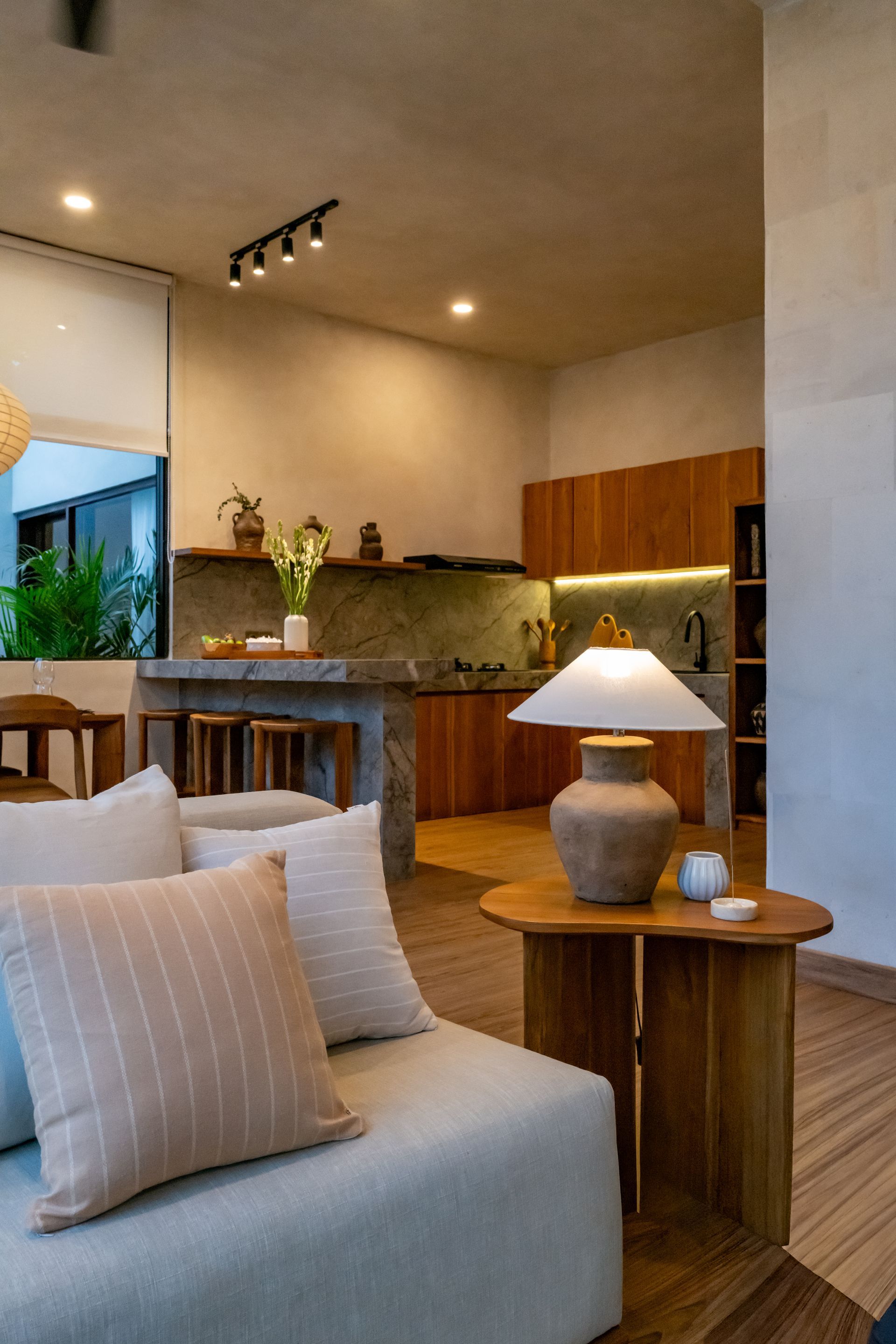 Cozy interior: Living area with light-colored sofa, wooden side table, lamp, and visible kitchen with wood cabinetry.