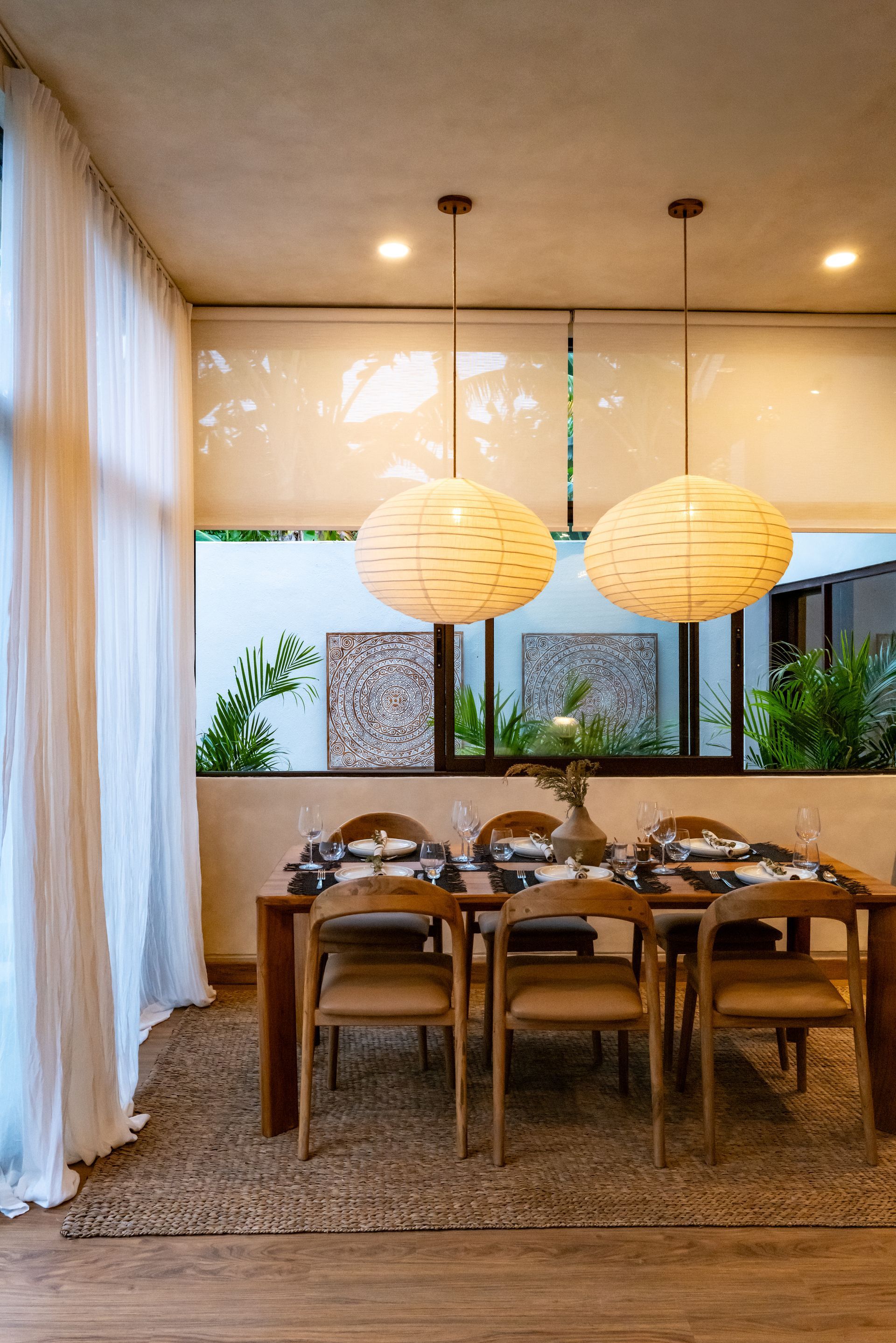 Dining room with wood table, chairs, and woven pendant lights. Sheer curtains and outdoor view.