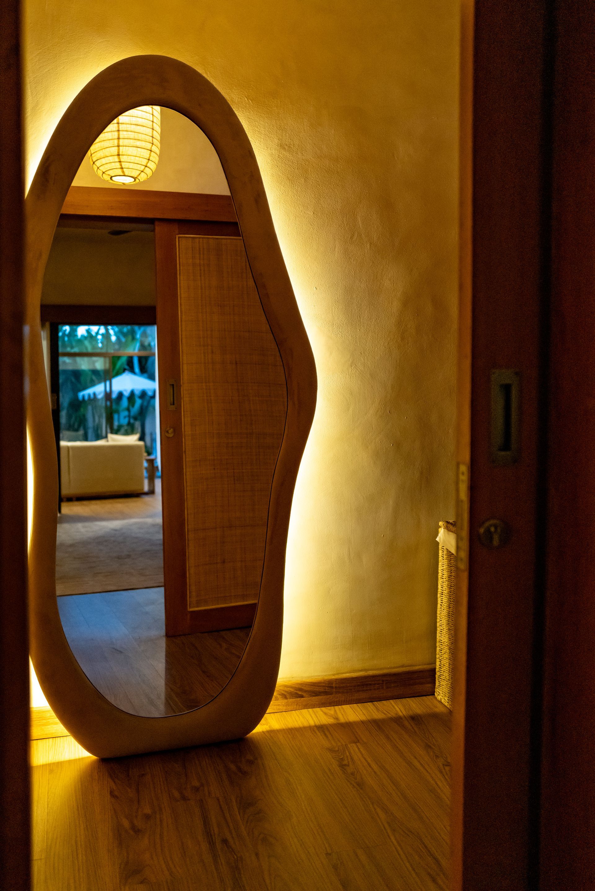 Large, organic-shaped mirror with built-in light in a room. Doorway view shows another room with outdoor view.