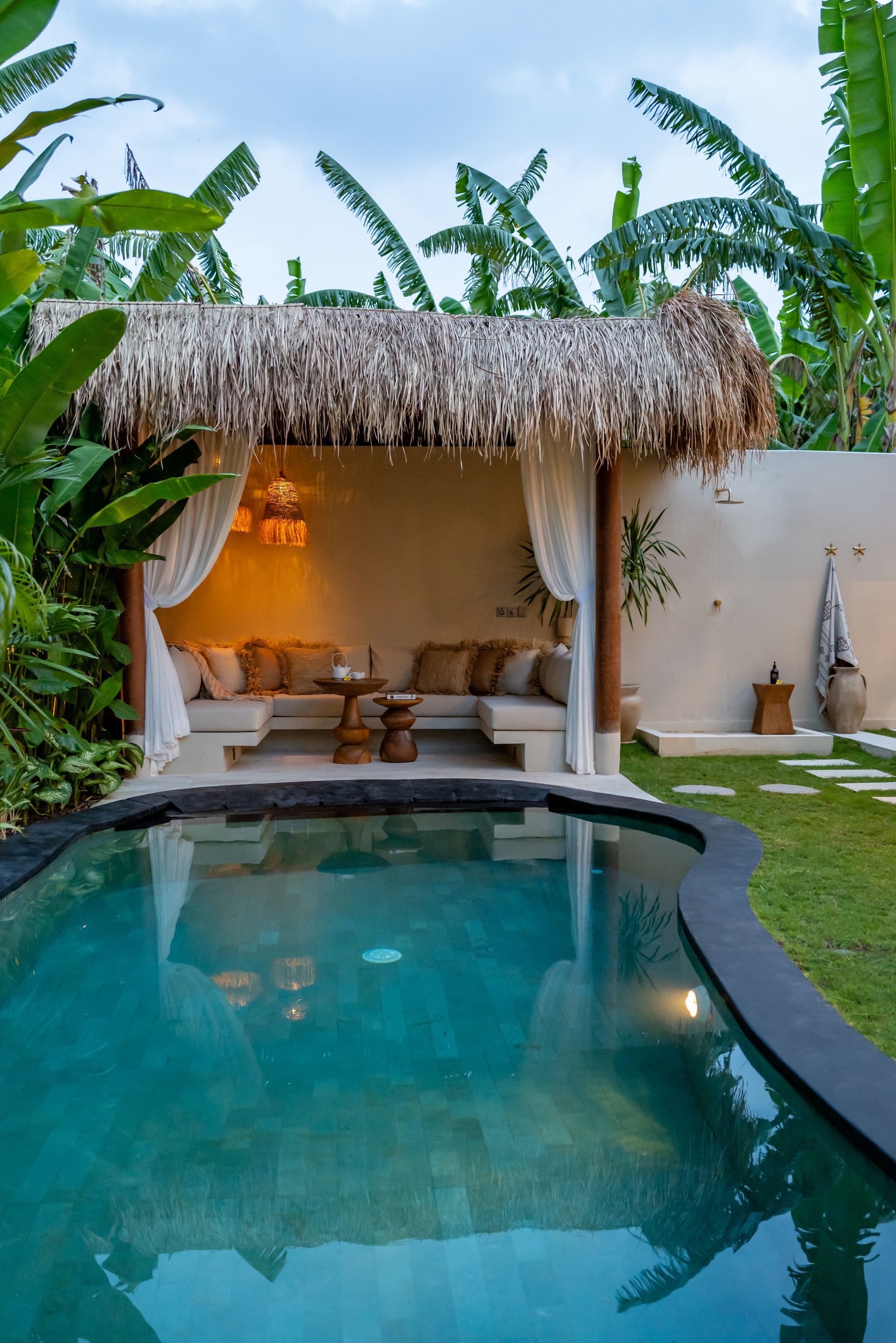 Poolside cabana with thatched roof, cushions, and sheer curtains. Turquoise pool surrounded by lush greenery.