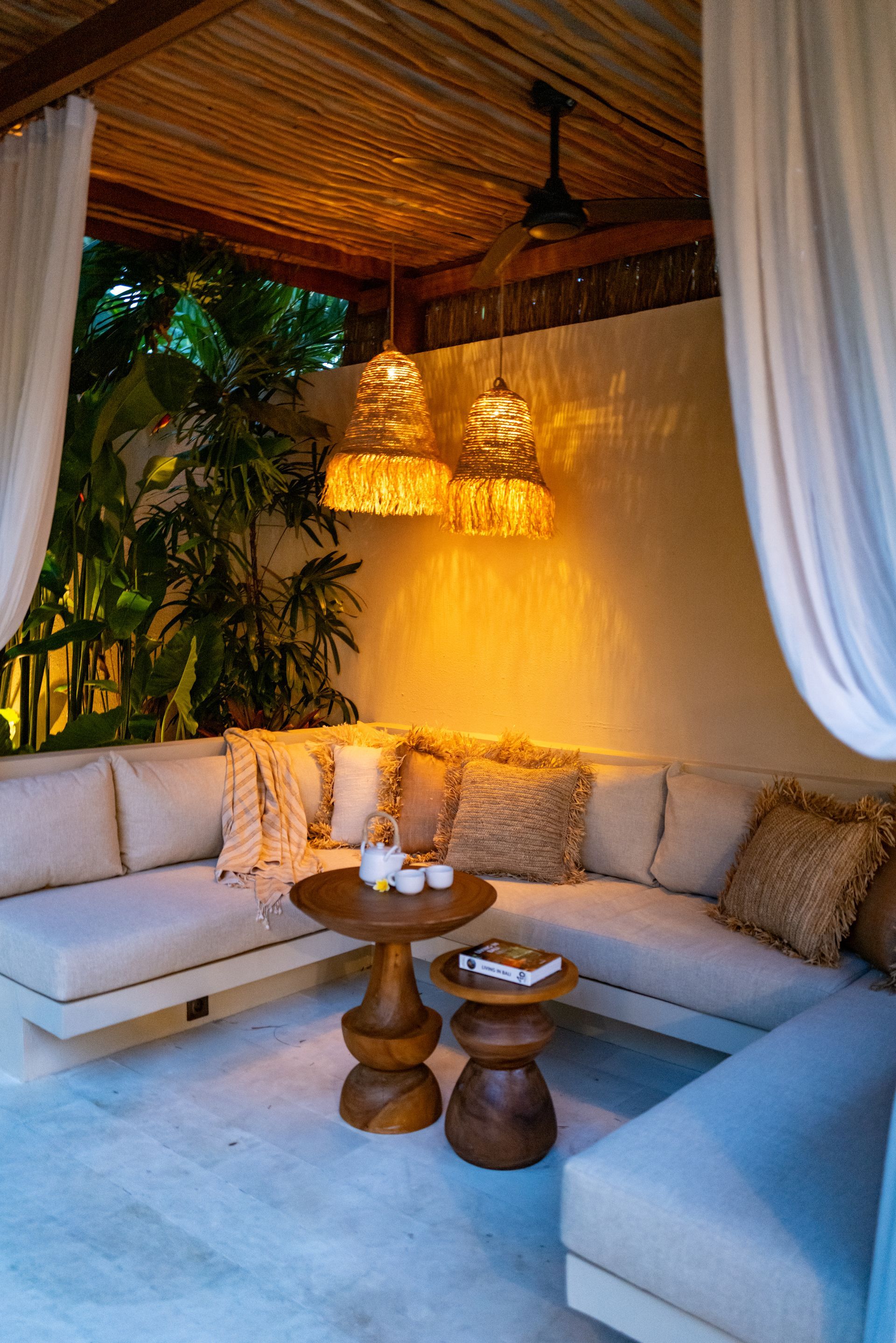 Cozy outdoor lounge area with woven lamps, cream-colored couches, wooden tables, and white curtains.