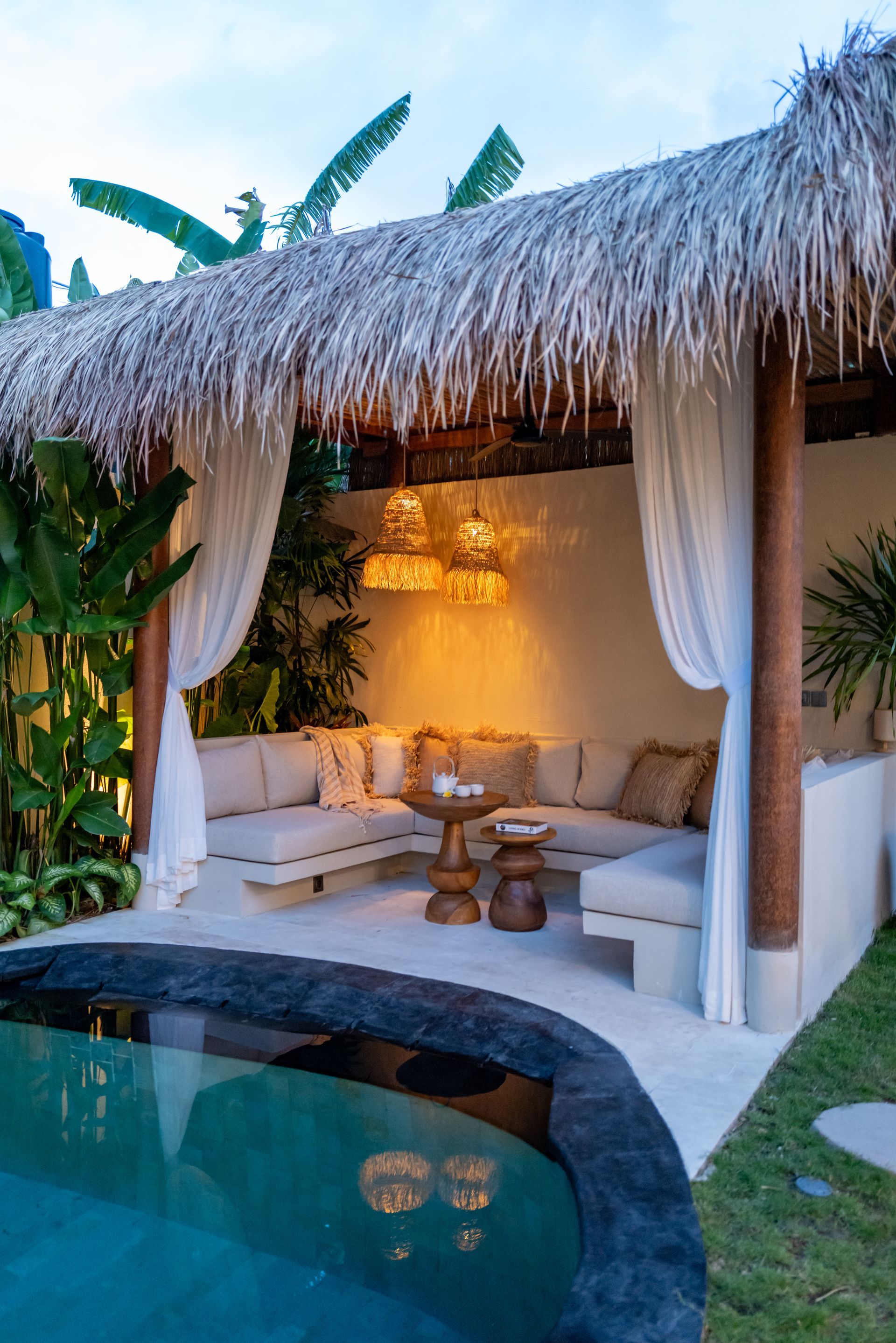 Tropical poolside lounge with thatched roof, cream seating, and hanging lights.