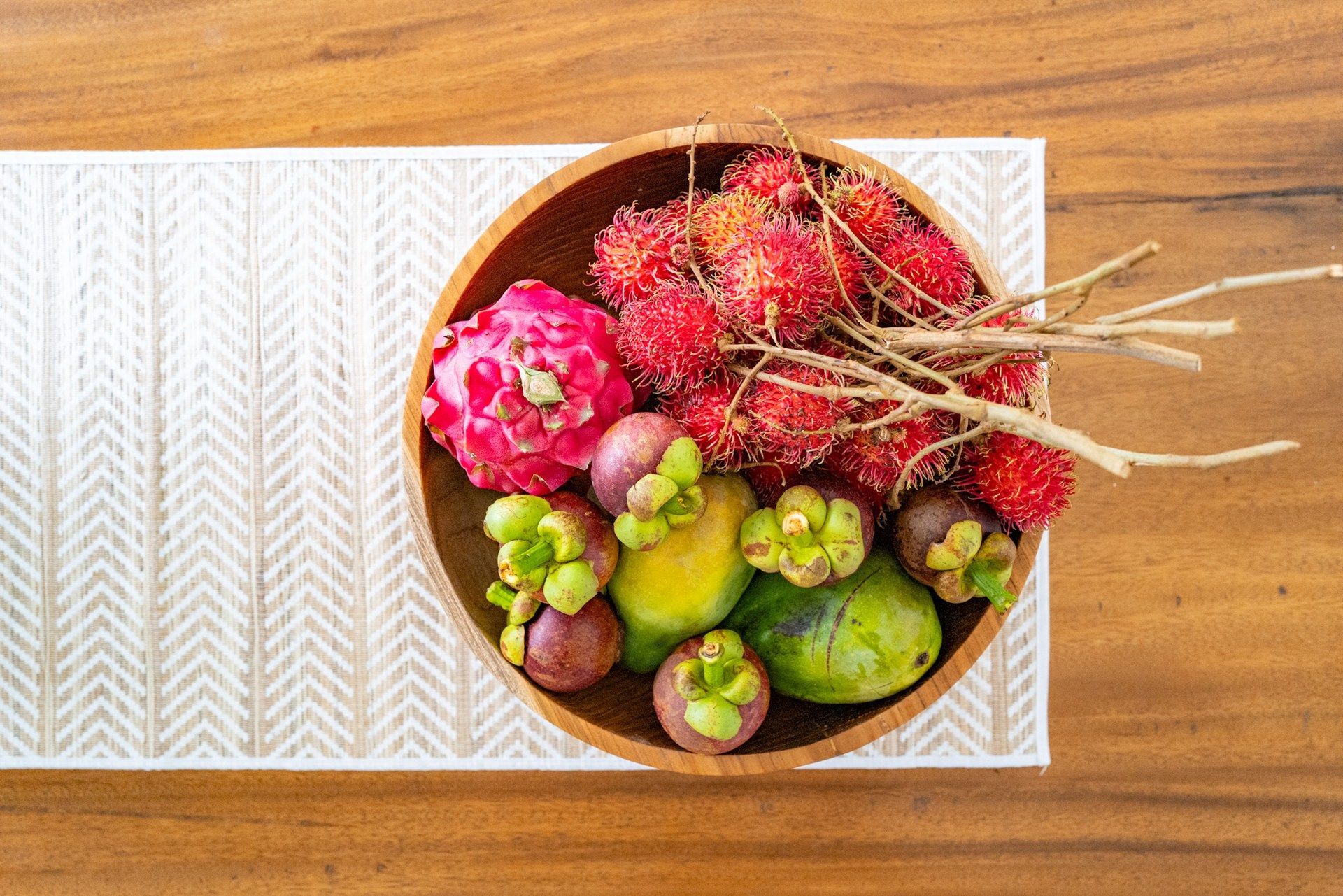 Wooden bowl filled with rambutan, mangosteen, mango, and a pink flower on a patterned runner on a wooden table.